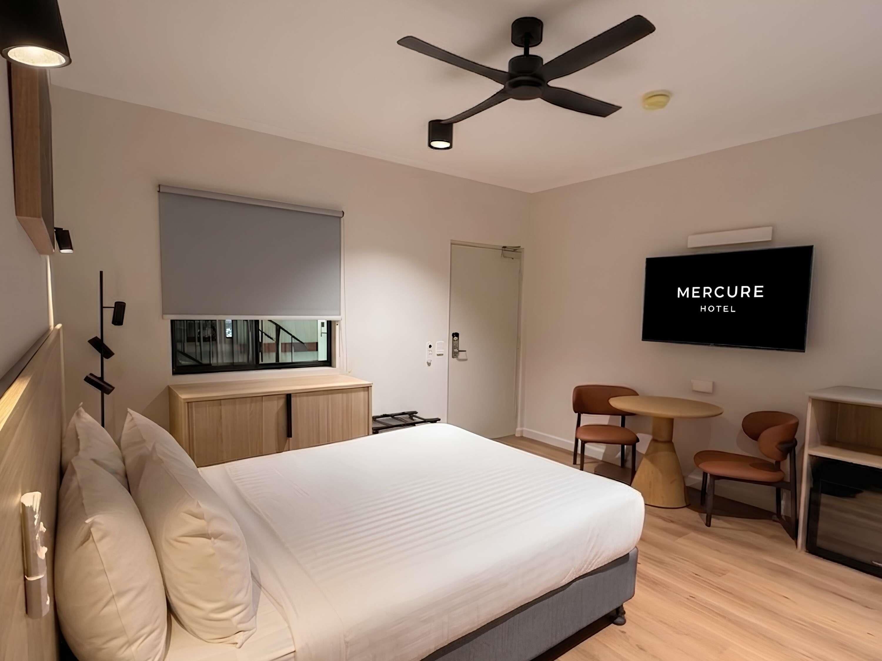 A modern hotel room with a bed, a ceiling fan, a TV, a desk, and a chair.