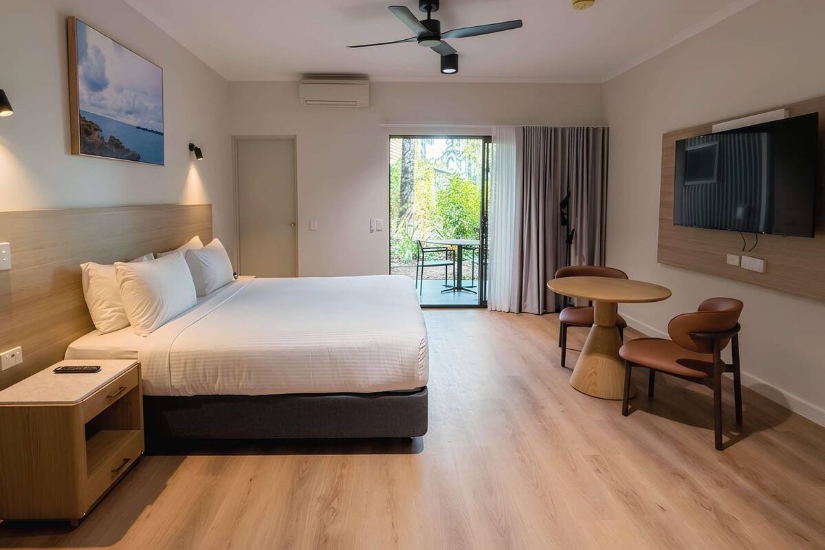 Quarto de hotel com cama, mesa de cabeceira, televisão e varanda com mesa e cadeiras.
