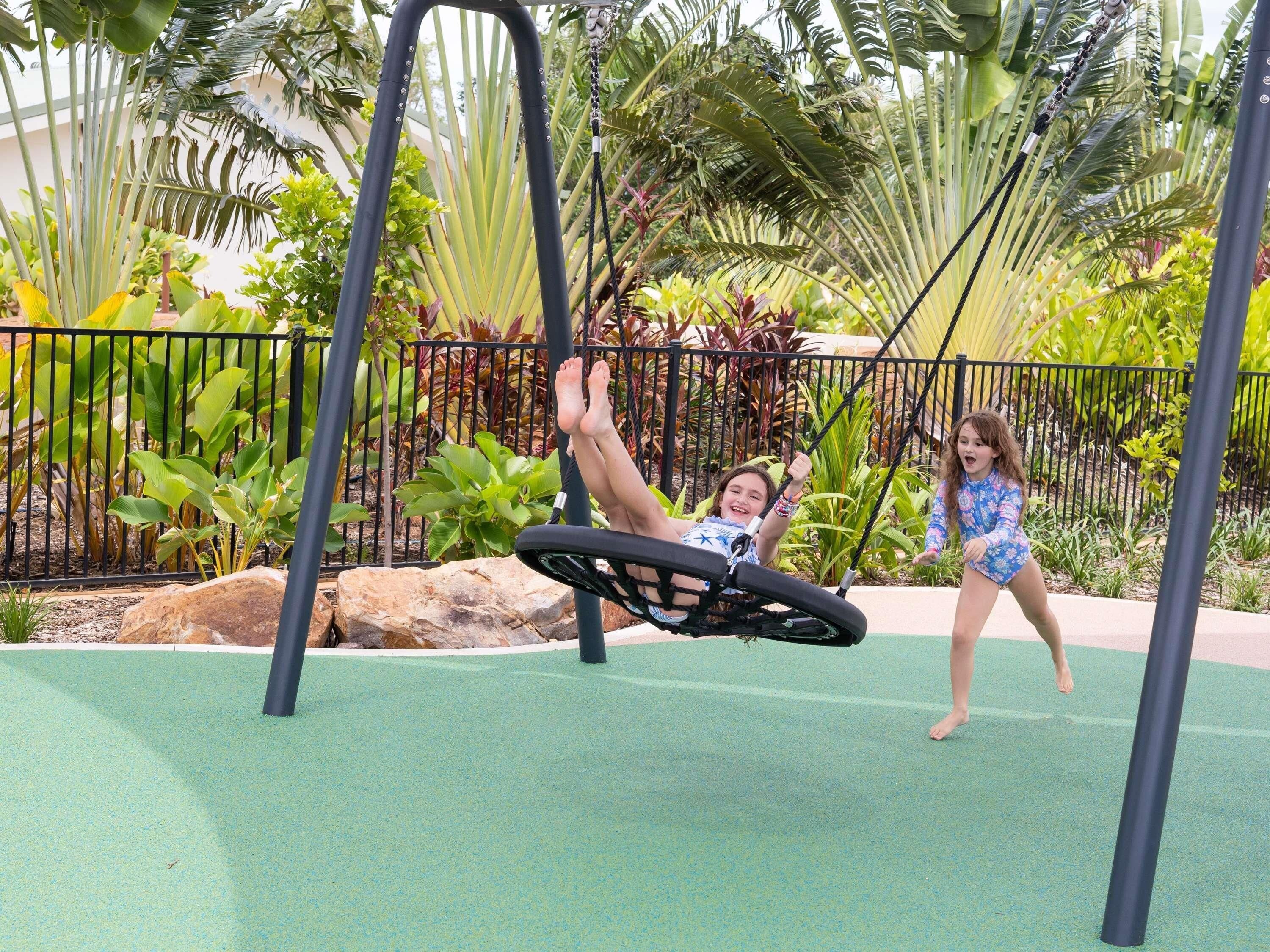 Un parque infantil con columpios y un área cercada con plantas tropicales.