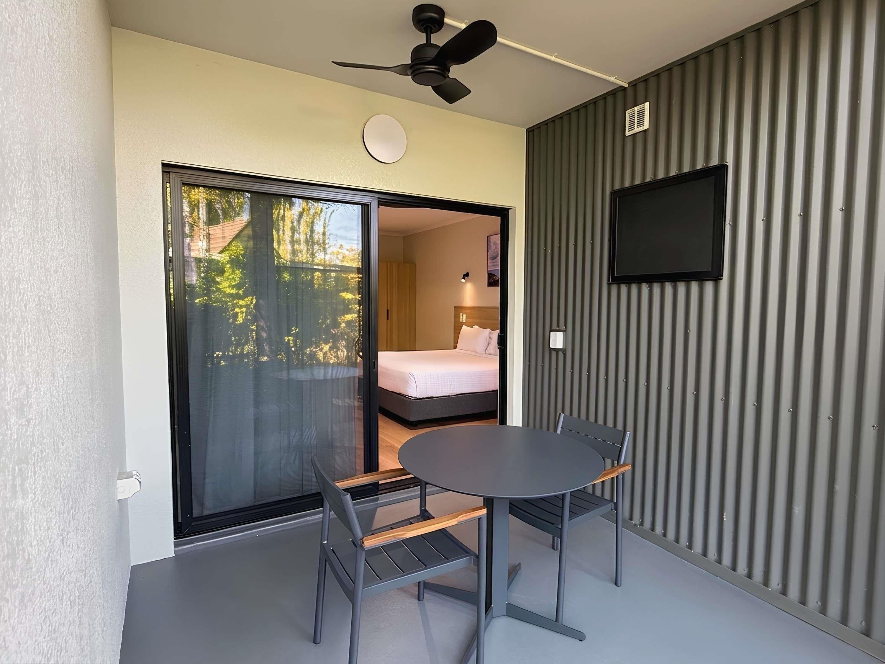 A room with a bed, a TV mounted on the wall, a ceiling fan, and a sliding glass door leading to an outdoor area.