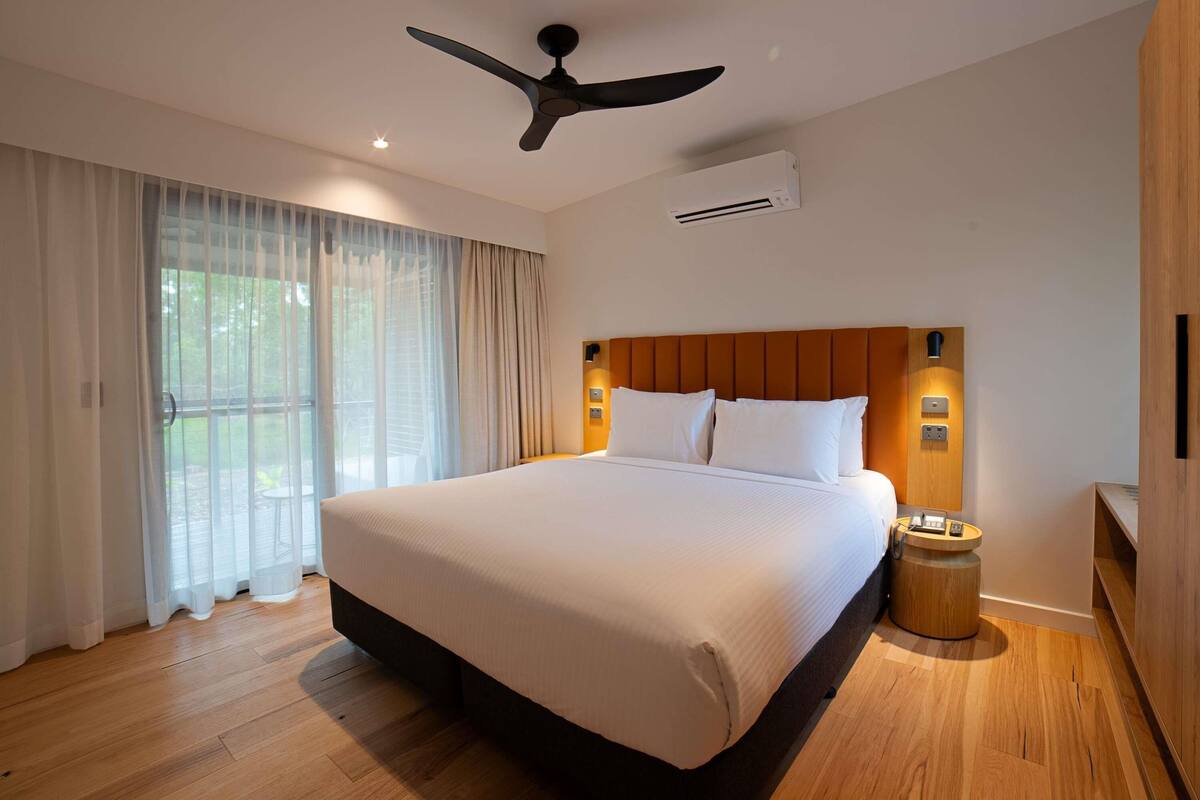 Quarto de hotel com uma cama grande, um ventilador de teto, um aparelho de ar condicionado, uma mesinha de cabeceira de madeira e vista para o exterior através de cortinas finas.