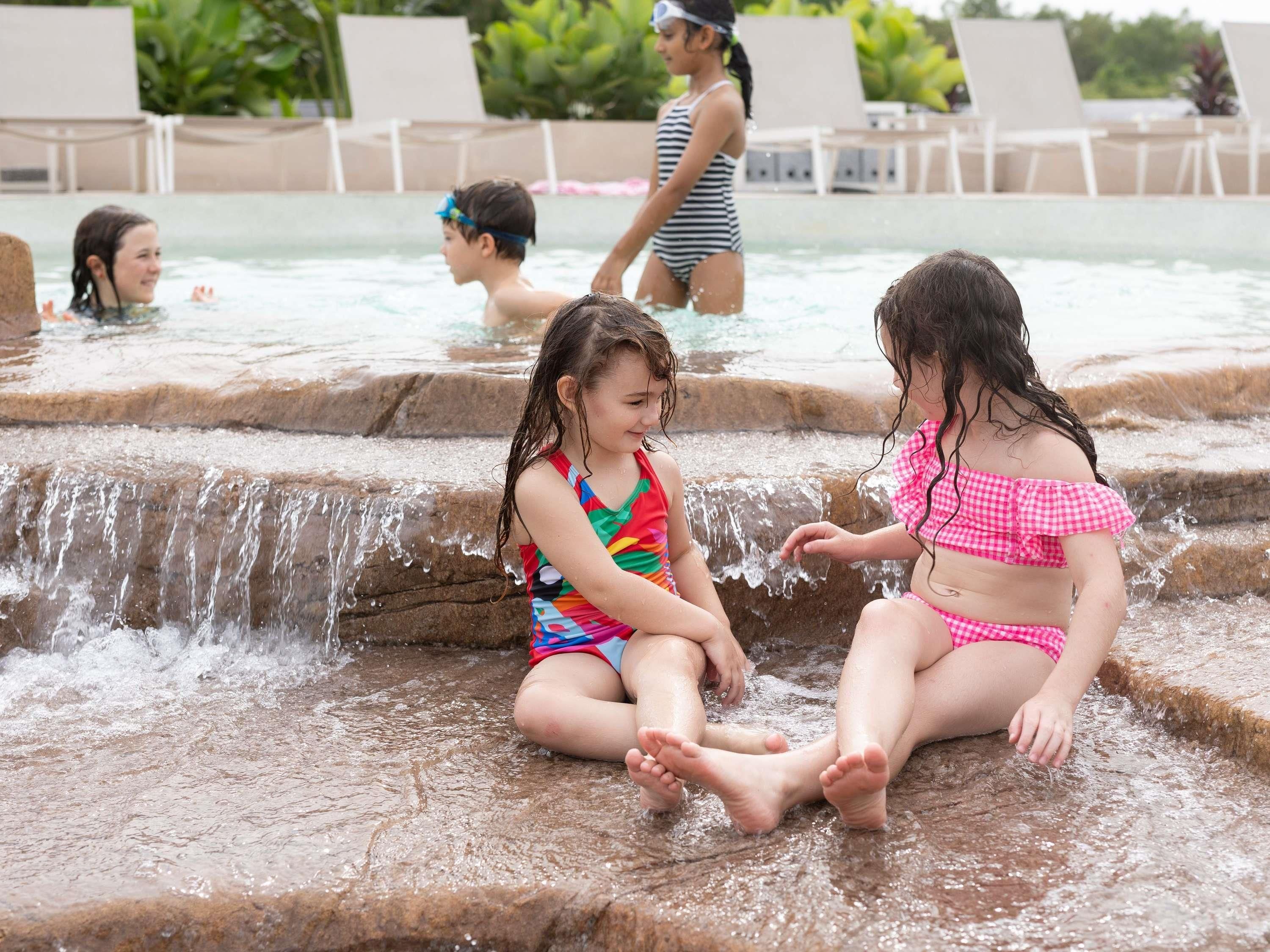 Crianças brincam em um espaço aquático, com piscina e cadeiras de descanso ao fundo.