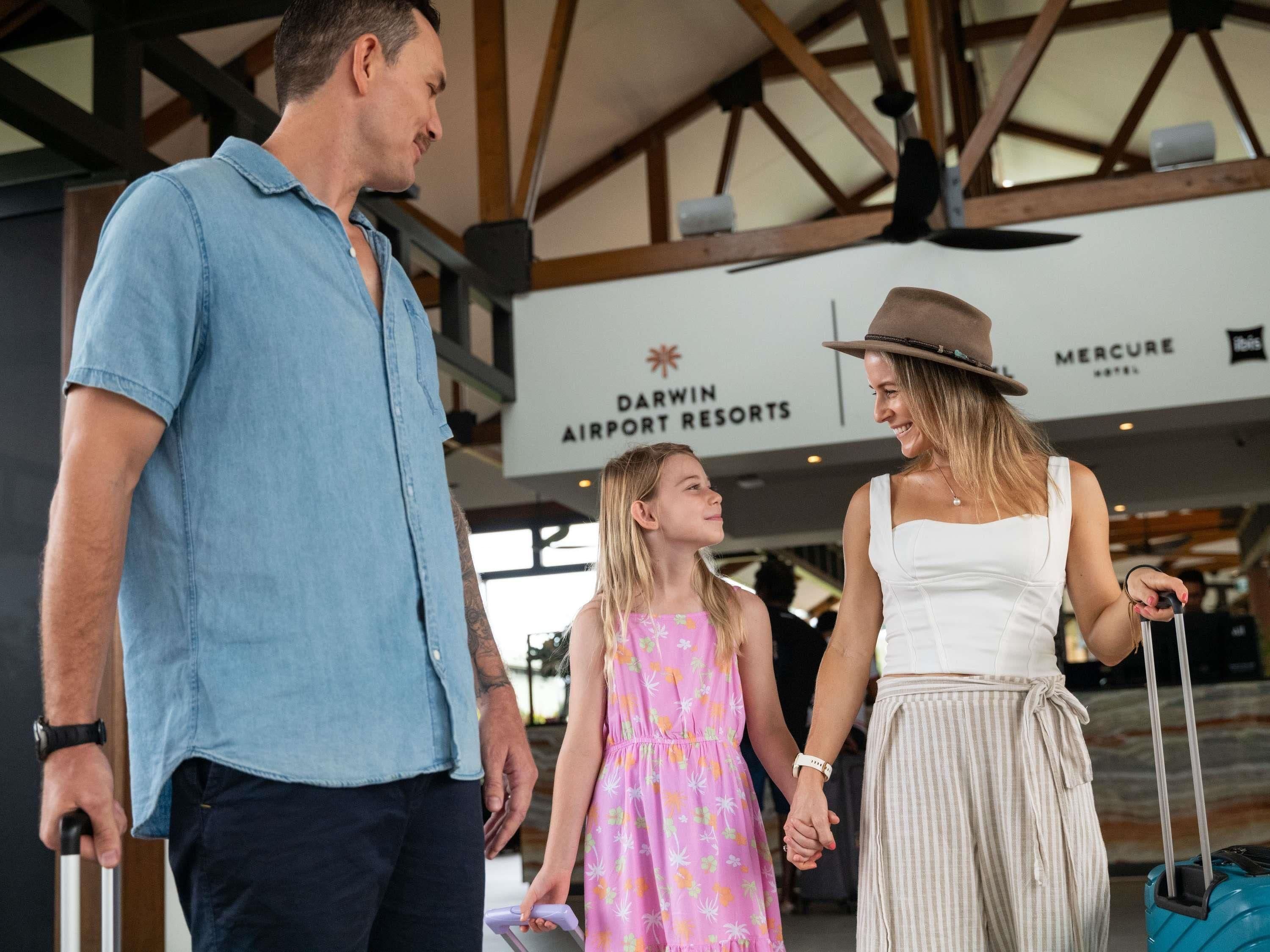 A family at an airport with a sign for Darwin Airport Resorts and a Mercure hotel in the background.