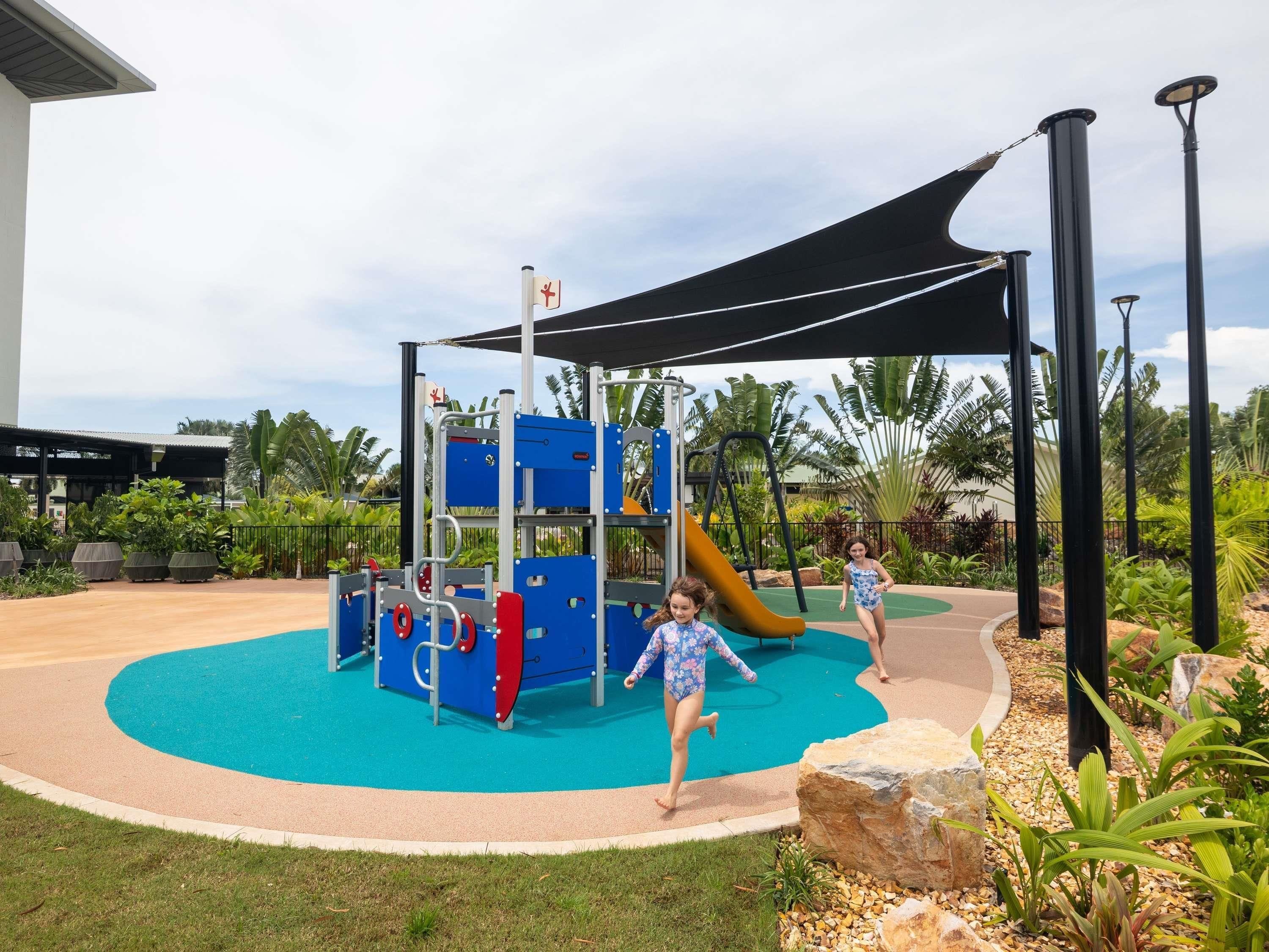 Un espace de jeux pour enfants avec un toboggan et des structures d’escalade, entouré d’un jardin paysager agrémenté de palmiers et d’un espace de repos abrité.