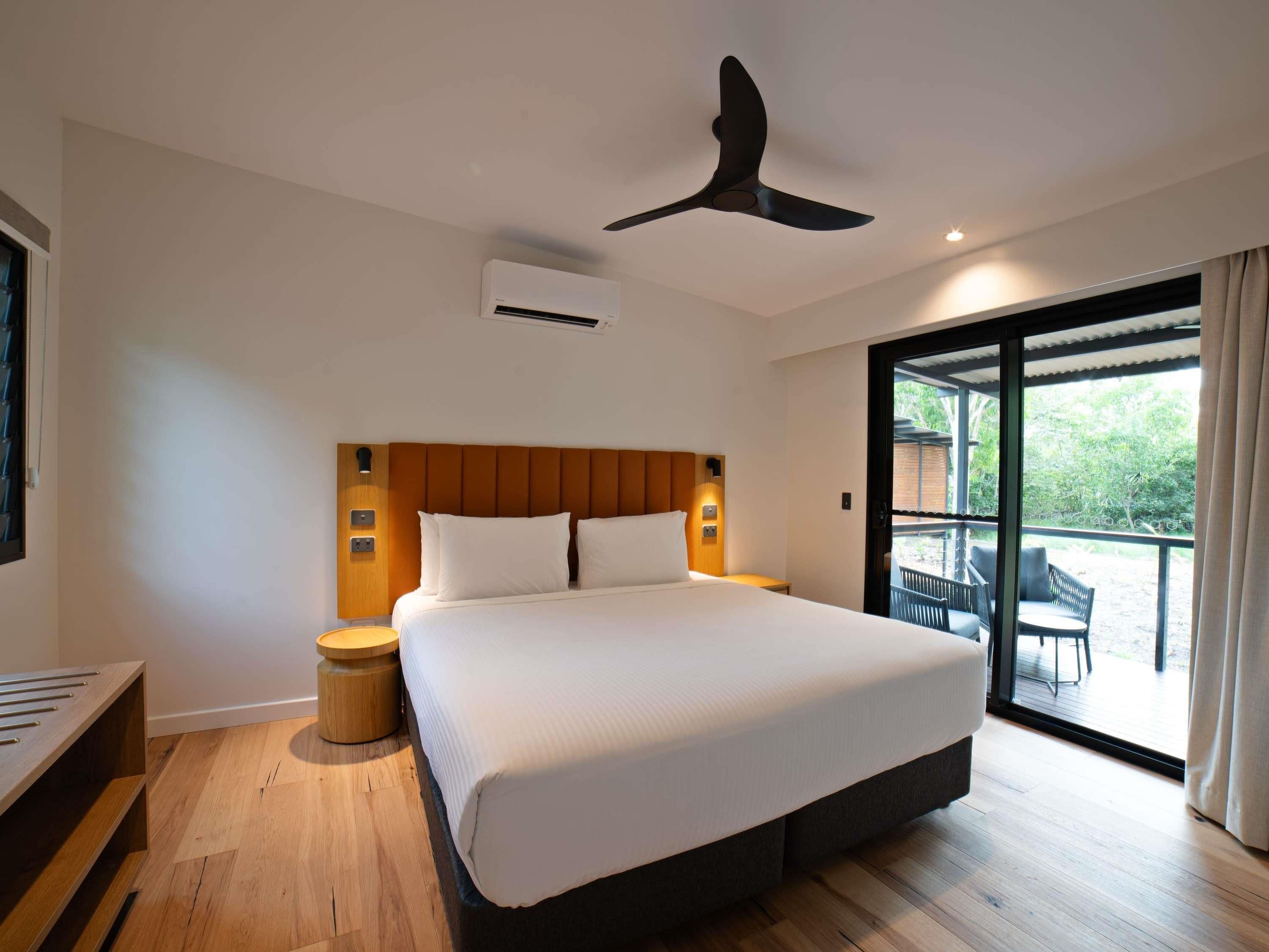 A modern hotel room with a large bed, a ceiling fan, an air conditioning unit, a wooden headboard, a small round table, and a view of the outdoors through a sliding glass door.