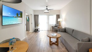 Habitación de hotel con piso de madera, un sofá gris, una mesa redonda para café, un televisor mostrando una escena de playa y un ventilador de techo.