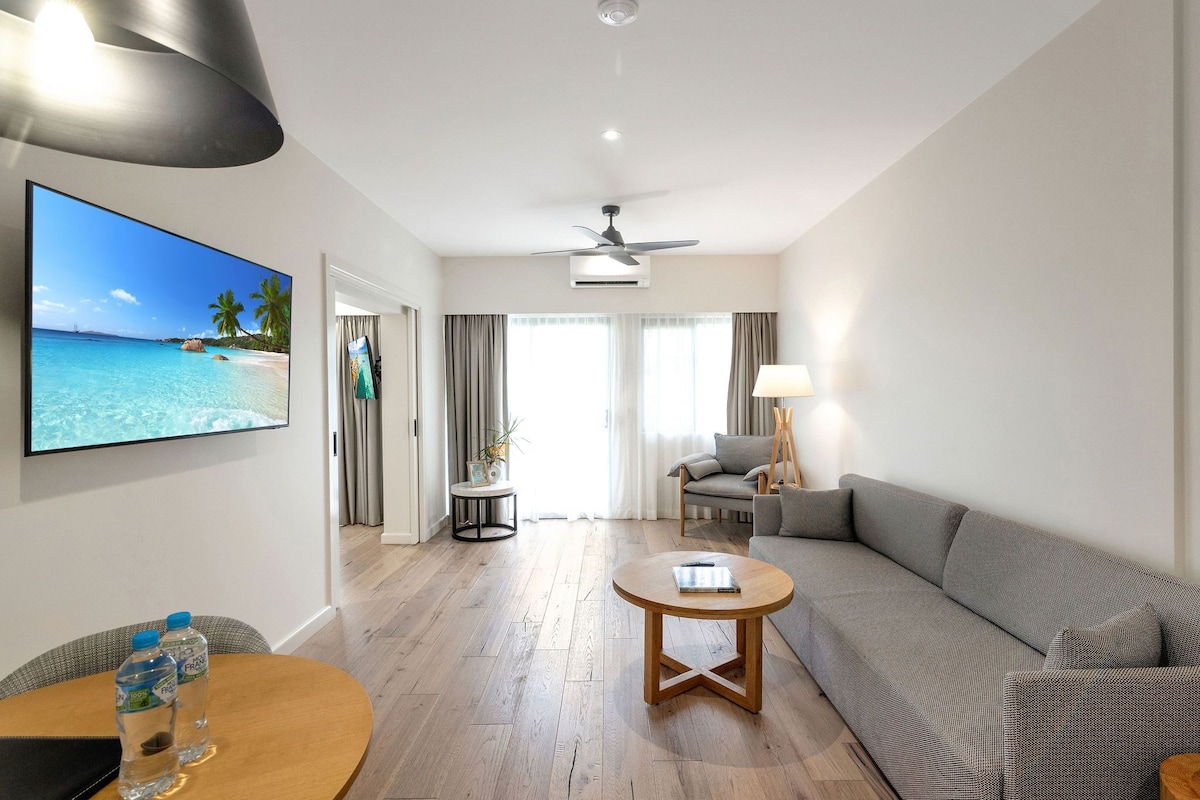 A hotel room with a wooden floor, a grey sofa, a round coffee table, a TV displaying a beach scene, and a ceiling fan.