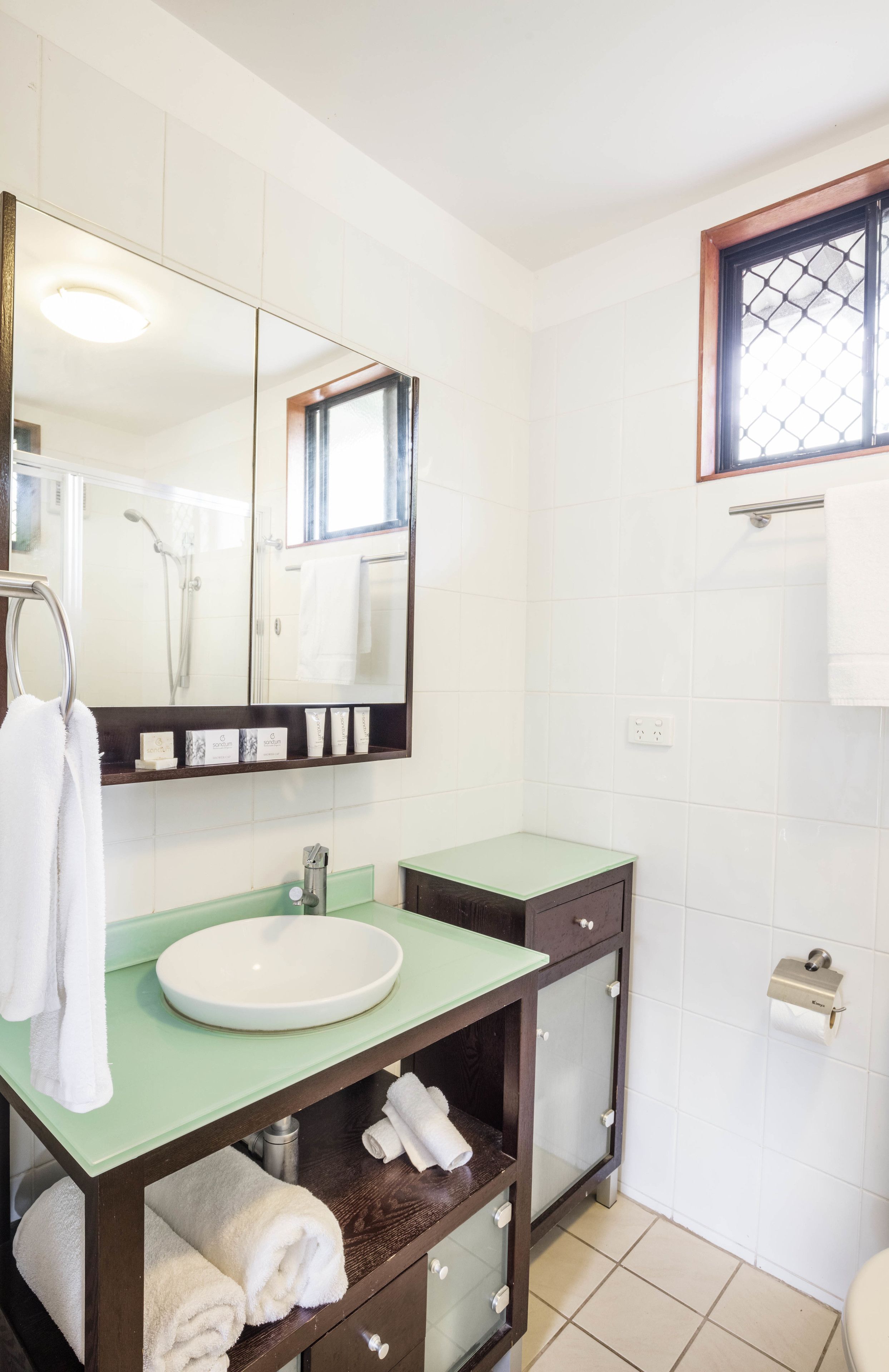 Un baño con un lavamanos blanco, encimera verde y un espejo grande. Hay un perchero con toallas, una zona de ducha y una ventana con celosía metálica.