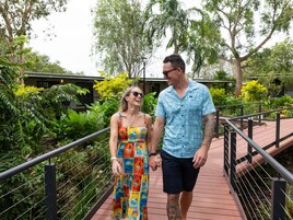 Um casal caminhando sobre uma ponte, com vegetação e um edifício ao fundo.