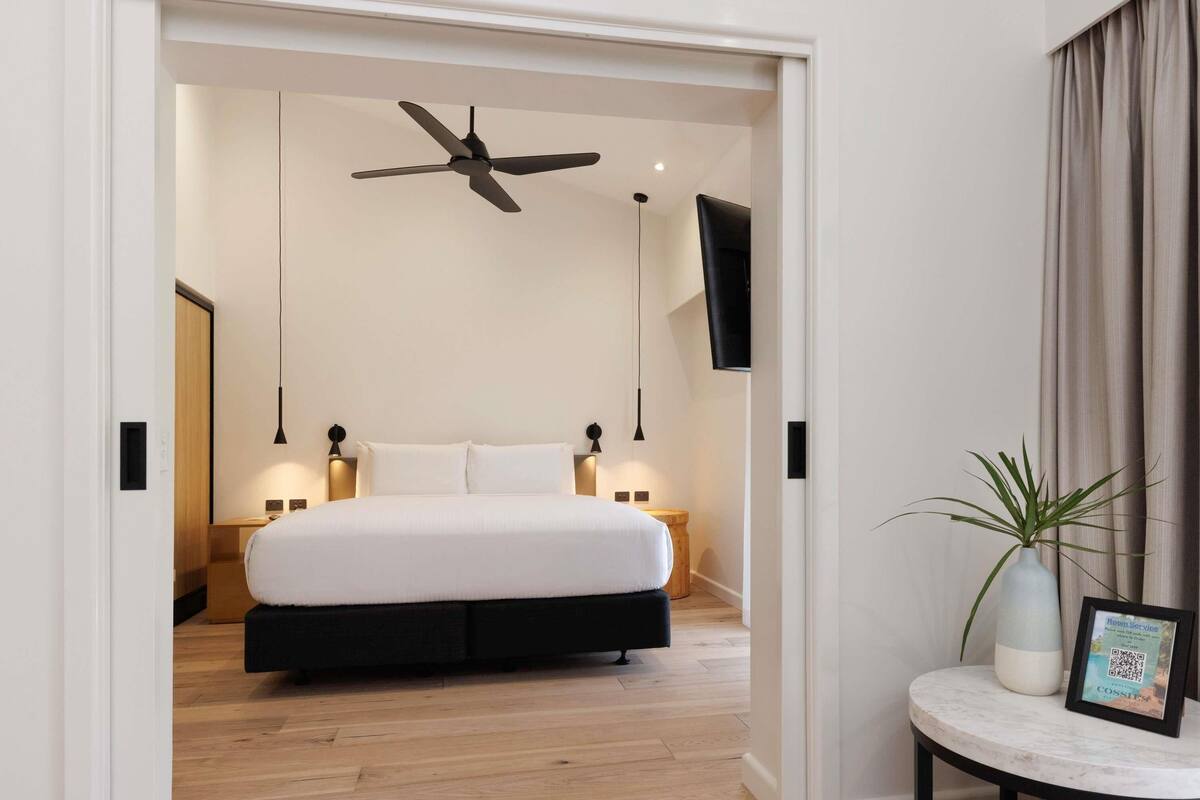 Quarto moderno com cama grande, ventilador de teto, televisão, abajures e uma pequena mesa com uma planta.