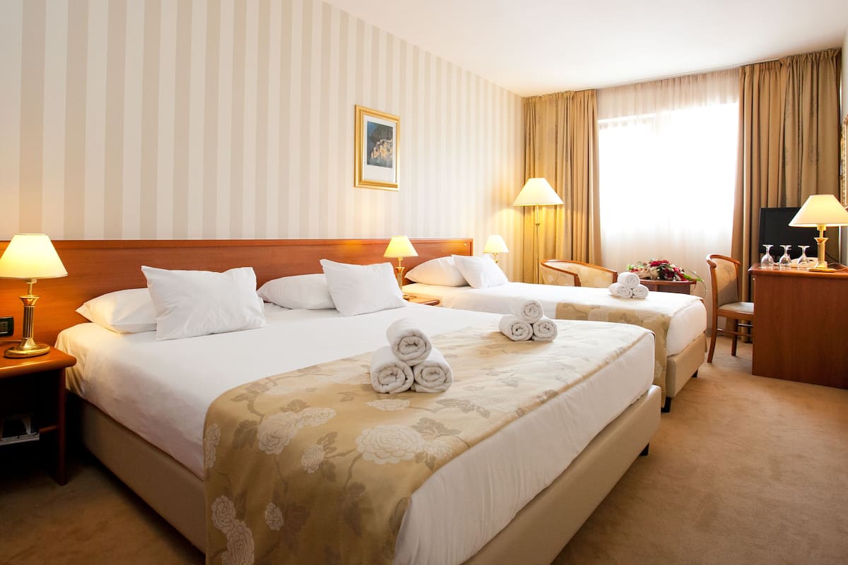 Quarto de hotel com duas camas, cabeceira de madeira, mesa de cabeceira com abajur, escrivaninha com abajur e uma cadeira.