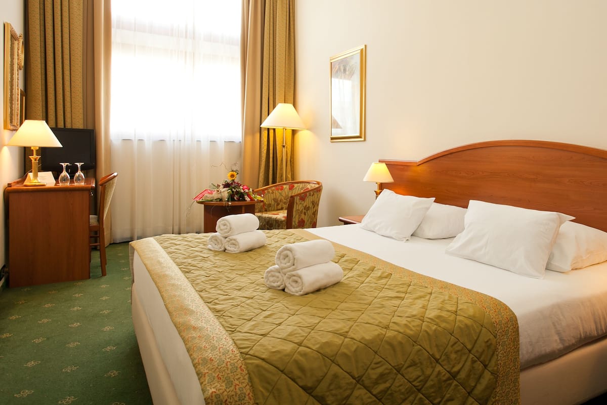 Quarto de hotel com uma cama grande, duas mesas de cabeceira com luminárias, uma cadeira, uma escrivaninha e uma televisão.