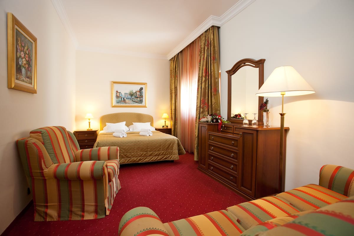 Quarto de hotel com uma cama, duas poltronas, uma cômoda e uma mesa de cabeceira com luminárias.