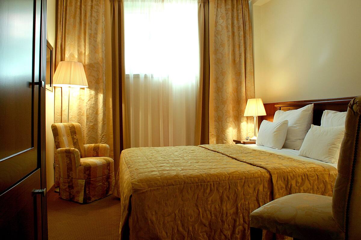 Quarto de hotel com uma cama, duas luminárias, uma cadeira e uma janela com cortinas.