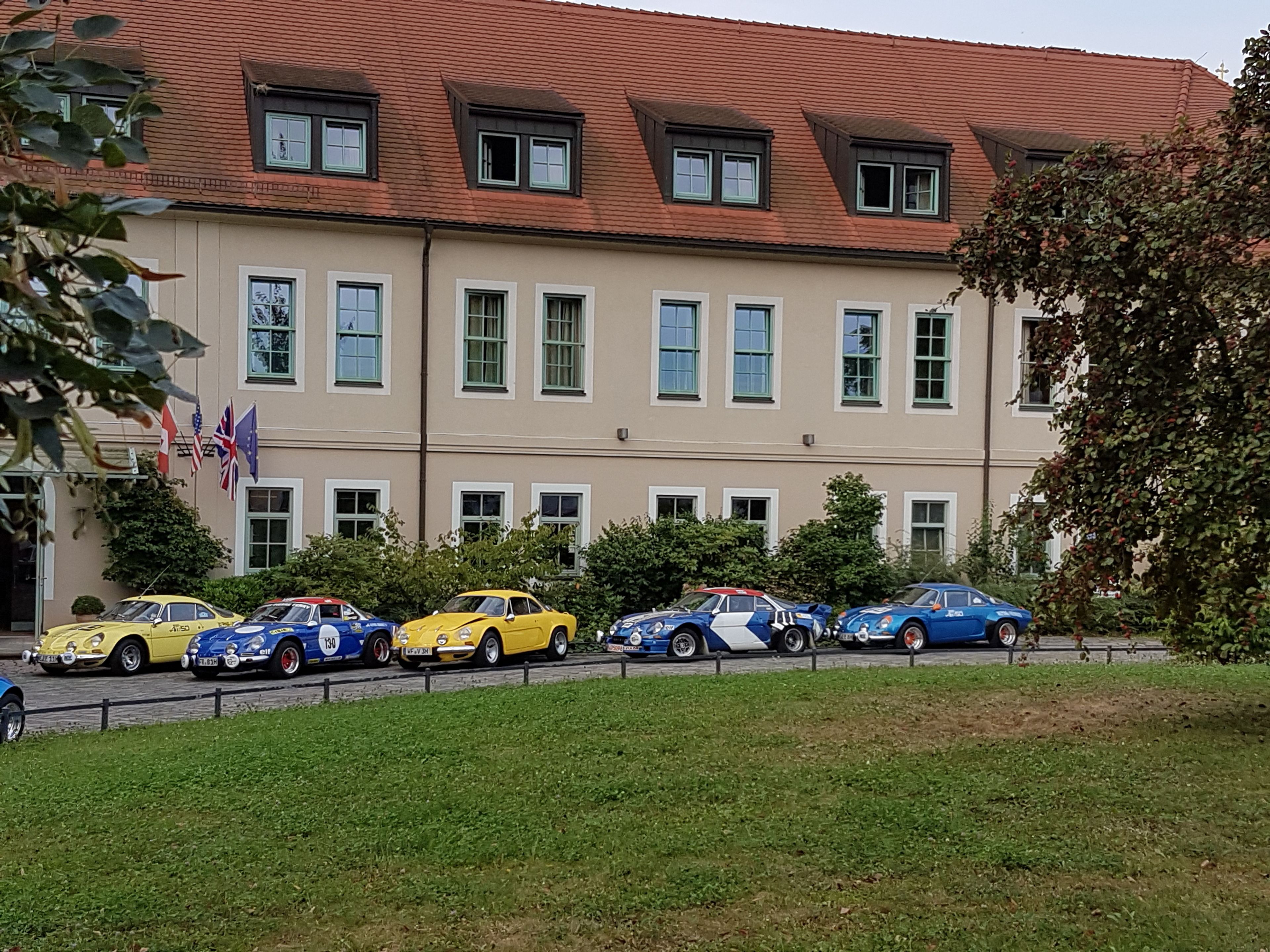 A row of classic cars parked in front of a building with a red-tiled roof and multiple windows.