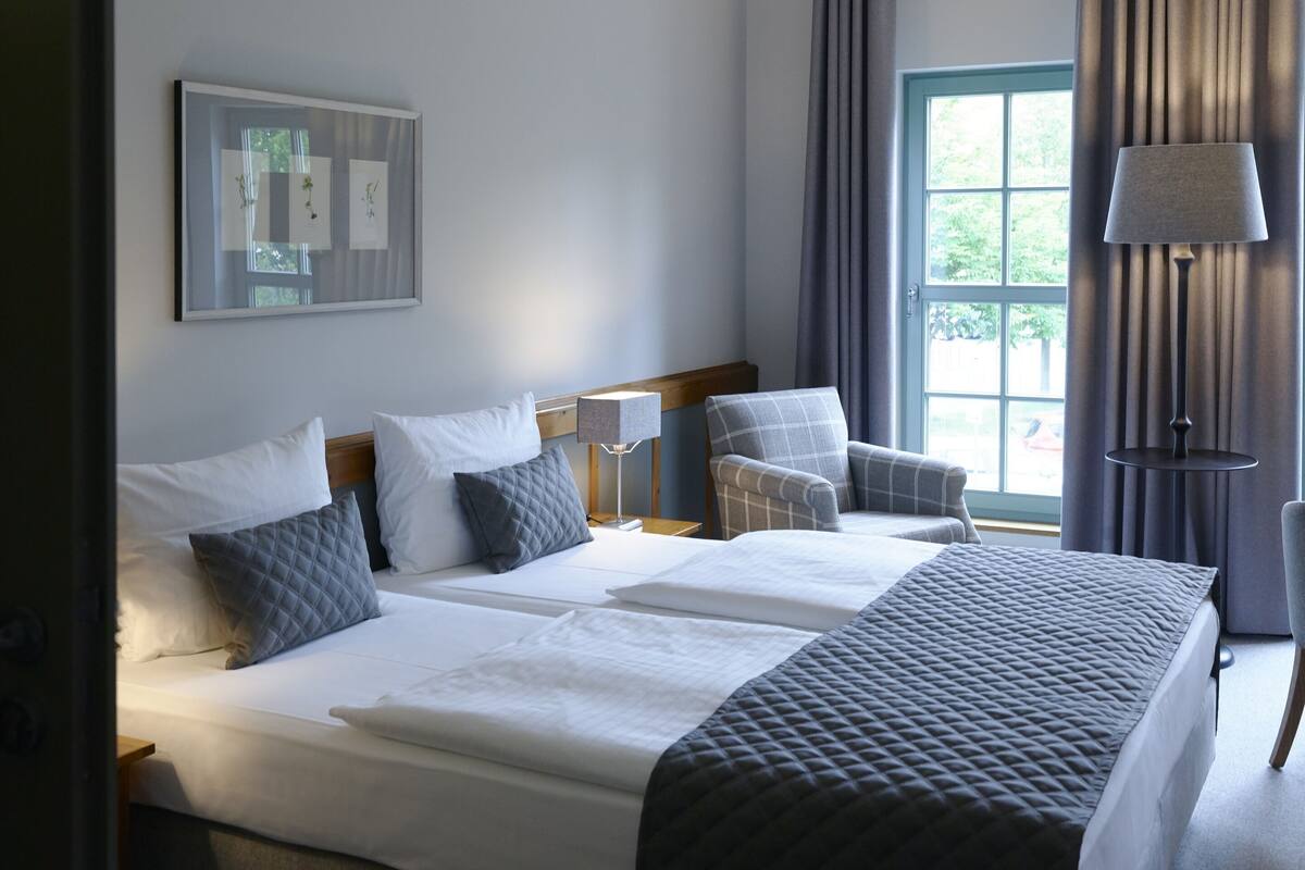 Ein Hotelzimmer mit einem großen Bett, einem Nachttisch, einer Lampe, einem Sessel und einem Fenster mit Vorhängen.