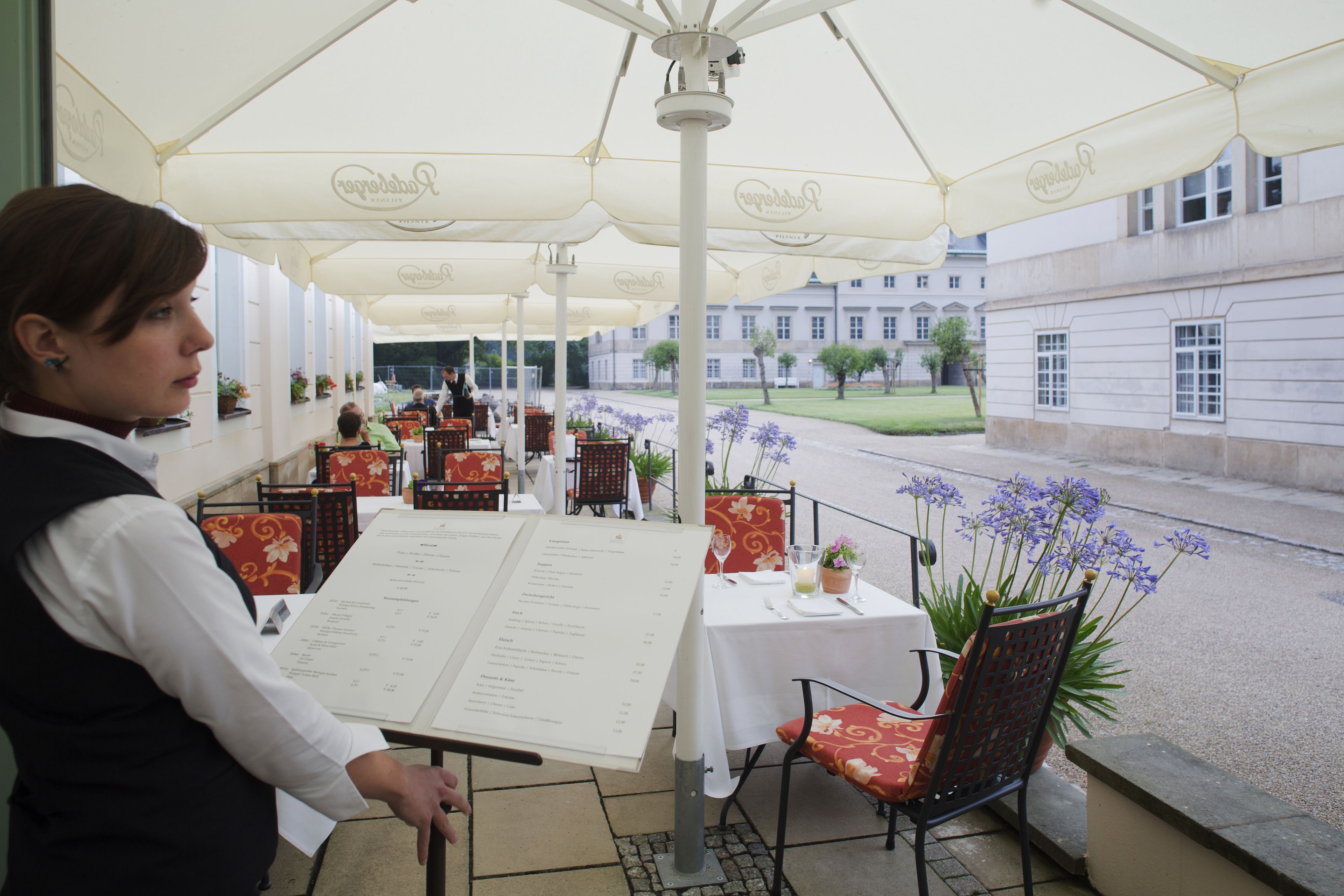 Un área de comedor al aire libre con mesas y sillas, un soporte para menús y vistas de un edificio al fondo.
