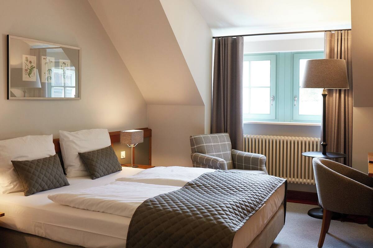 Ein Hotelzimmer mit einem Bett, zwei Stühlen, einem Schreibtisch, einer Lampe und einem fenster mit Vorhängen.