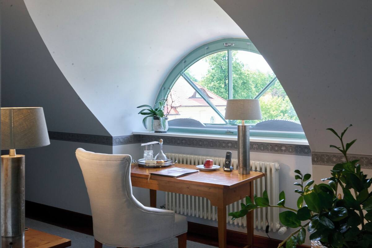 Ein Zimmer mit schrägem Dach, einem Holztisch mit Stuhl, einer Lampe, einem Blumentopf und Blick durch ein Fenster.