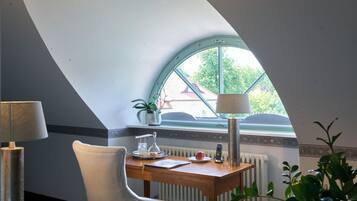Een kamer met een schuine hemel, een houten bureau met stoel, een lamp, een plant in pot en uitzicht door een raam.