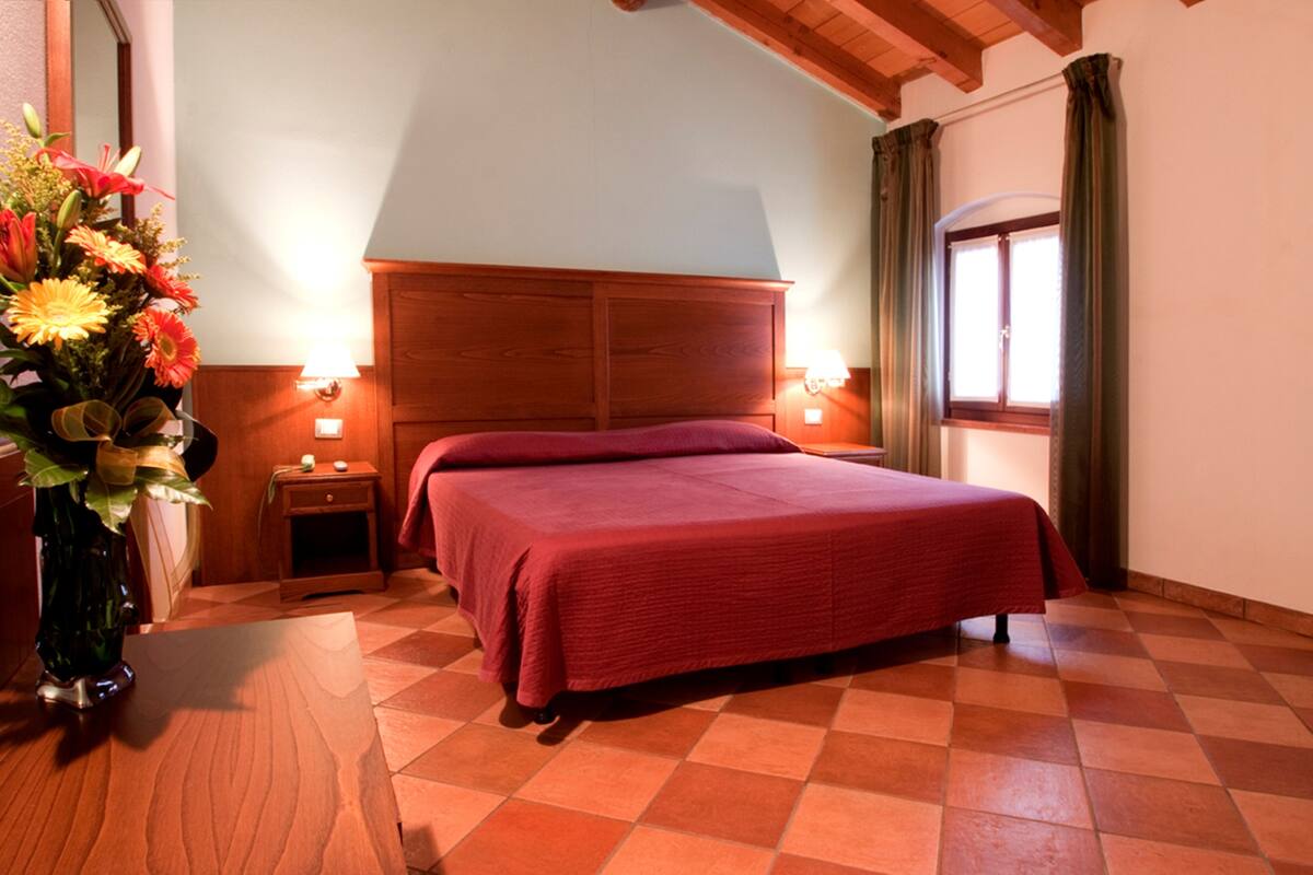 Une chambre à coucher avec une tête de lit en bois, un sol à carreaux, un lit recouvert d’une couverture rouge, une table de chevet avec une lampe et un vase rempli de fleurs.
