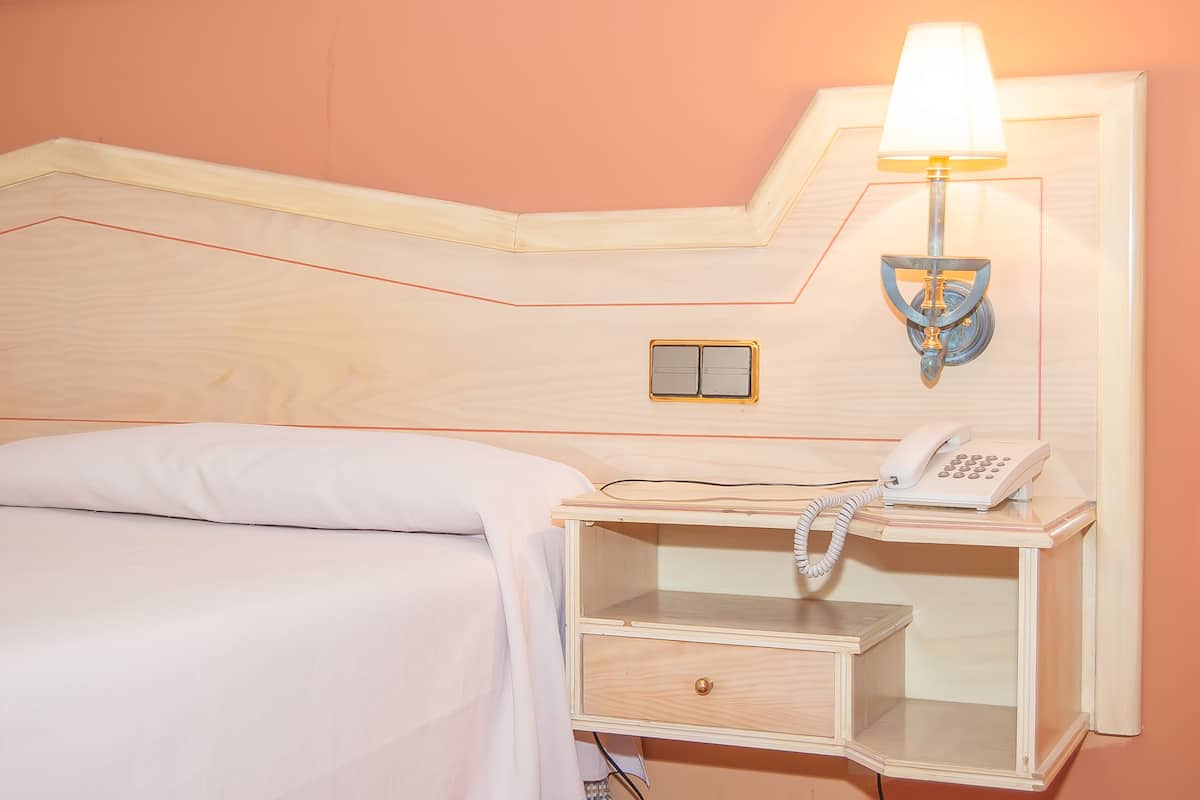 A neatly made bed with a white comforter and pillows, a bedside table with a lamp, a phone, and a small drawer.