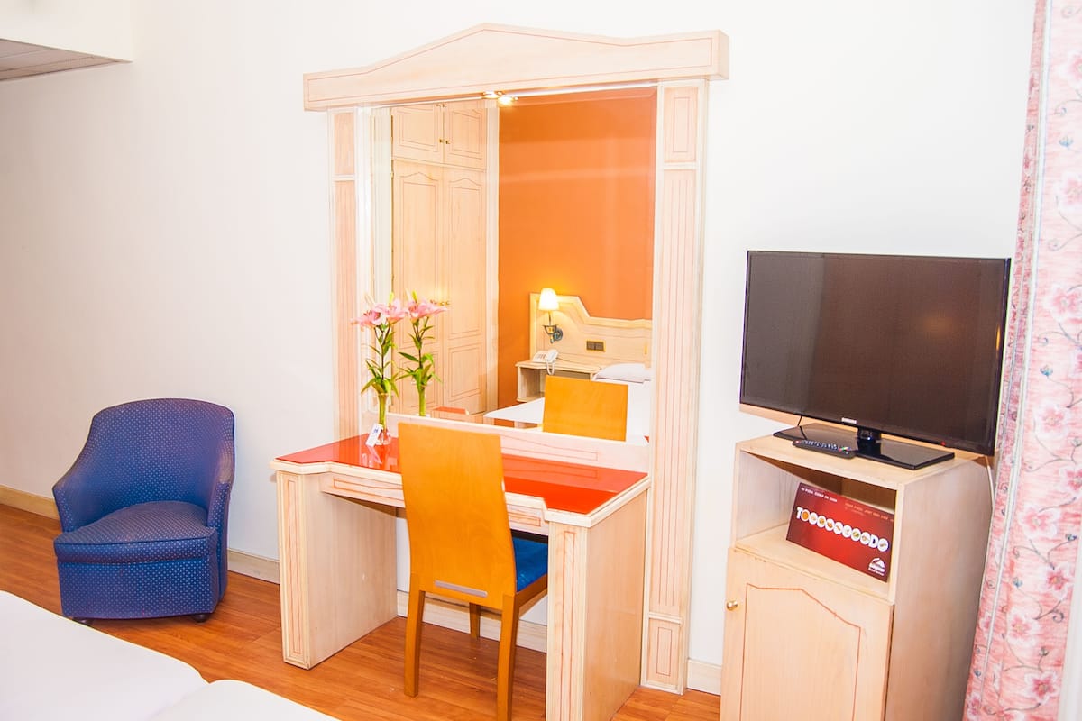 A hotel room with a wooden desk, a chair, a television, and a small cabinet.