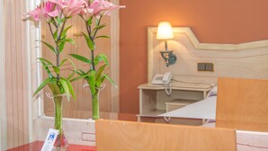 Una habitación de hotel con cabecera de madera, lámpara de noche, teléfono y un jarrón con flores rosas sobre una mesa.