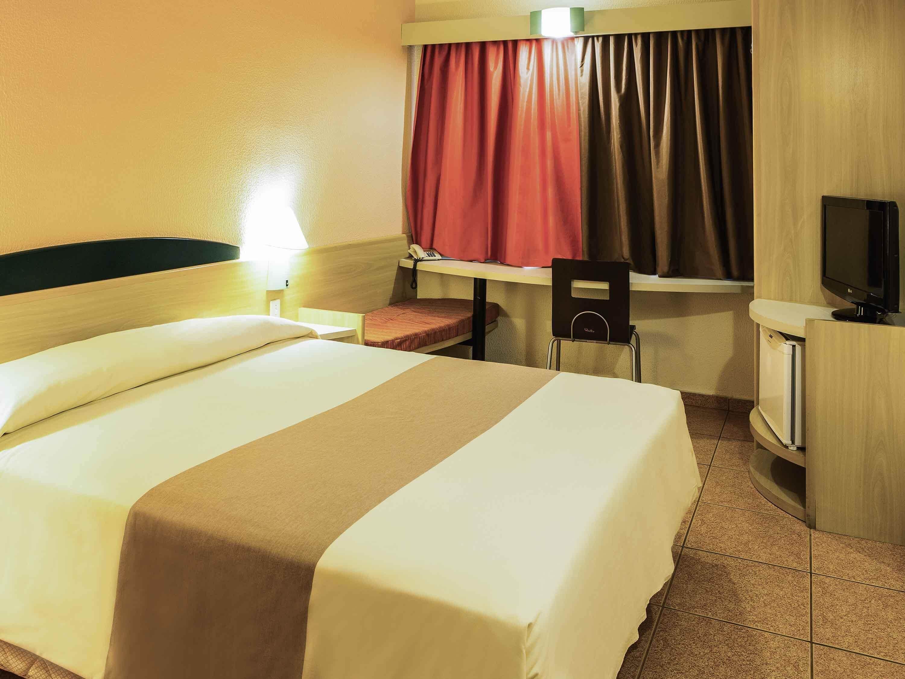 Een hotelkamer met een bed, een bureau, een stoel, een televisie en een raam met gordijnen.