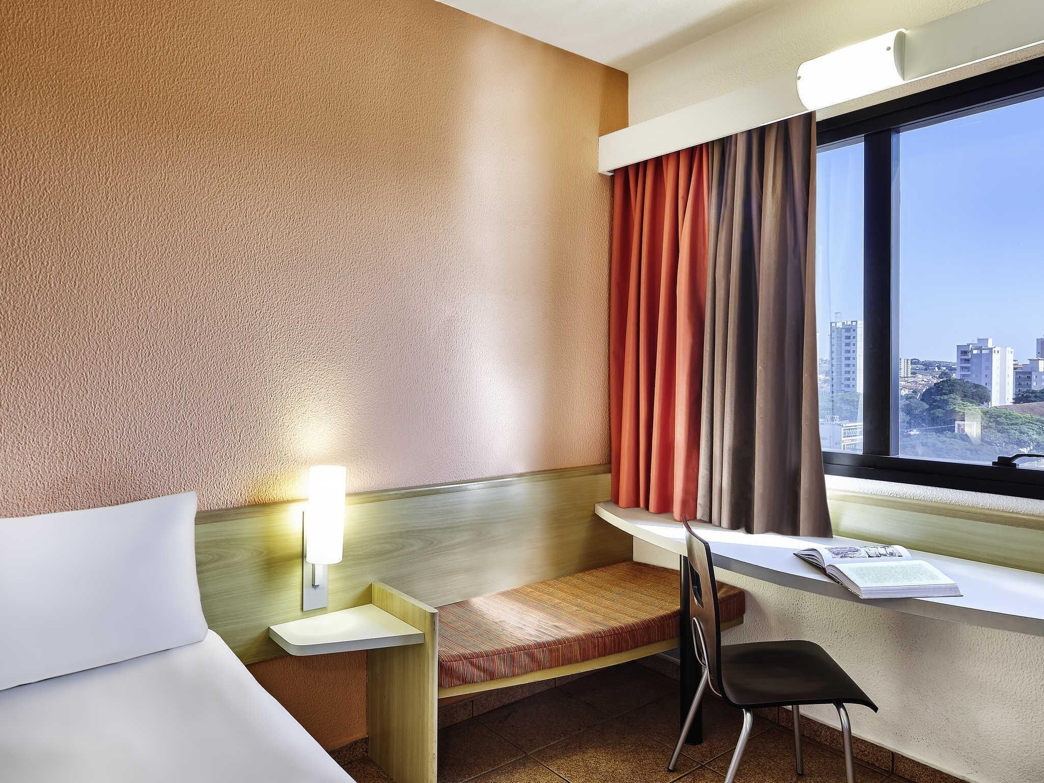 A hotel room with a bed, a desk with a chair, a window with curtains, and a city view.