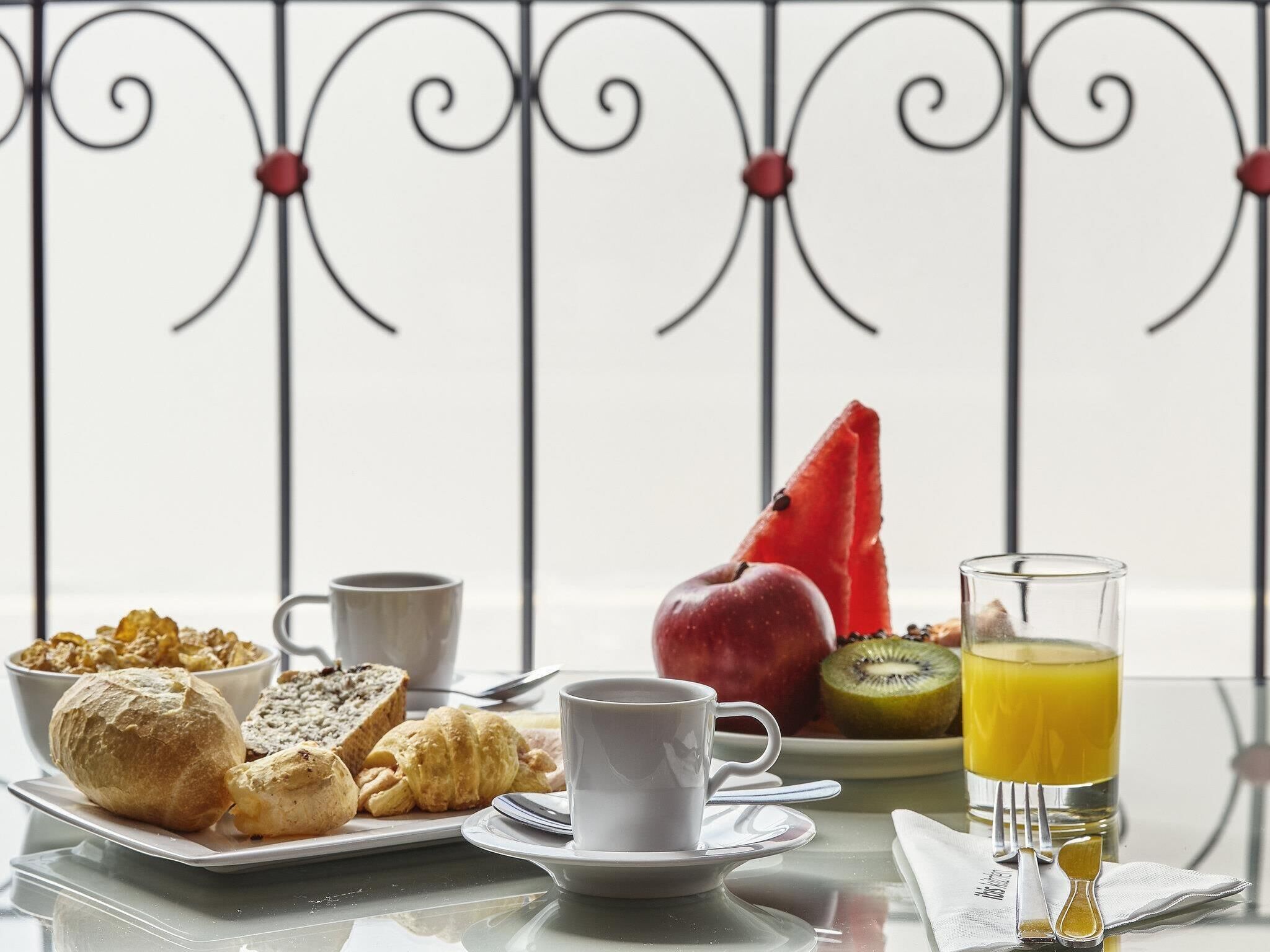 Une table dressée pour le petit-déjeuner, avec des viennoiseries, des fruits, une tasse et un verre de jus d’orange.