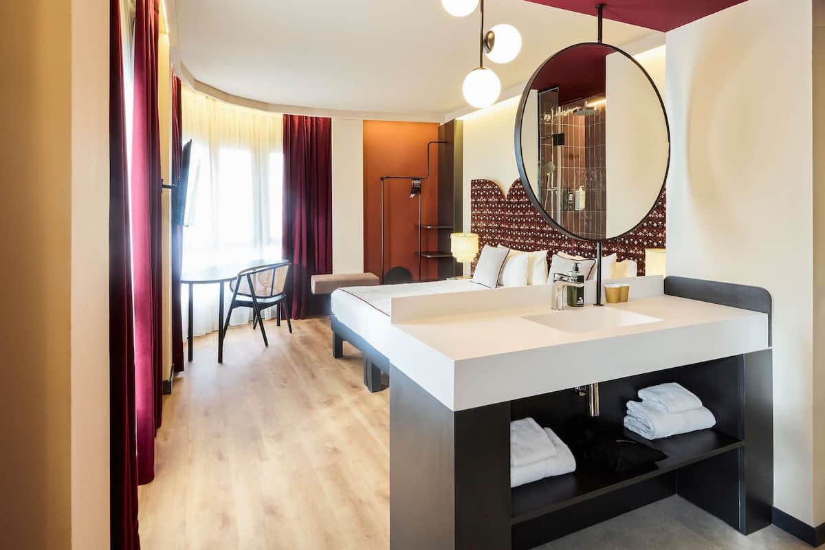 A modern hotel room with a large bed, a round mirror, a desk, and a chair.