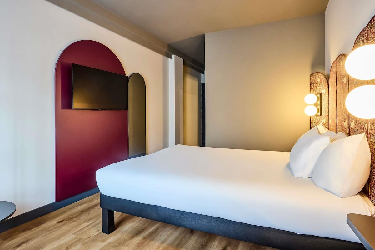 A modern hotel room with a large bed, a flat-screen TV mounted on a red wall, and a wooden floor.