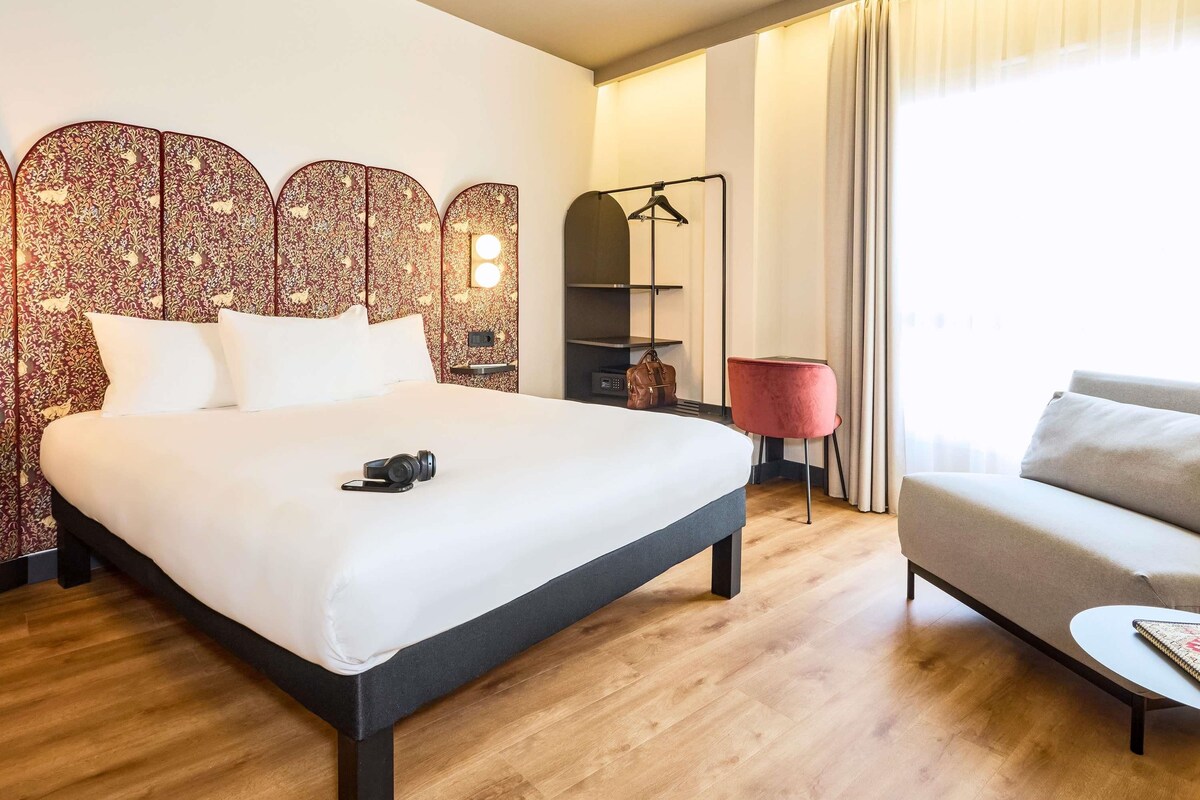 A hotel room with a bed, a sofa, a red chair, a hanging coat rack, and a mirror.