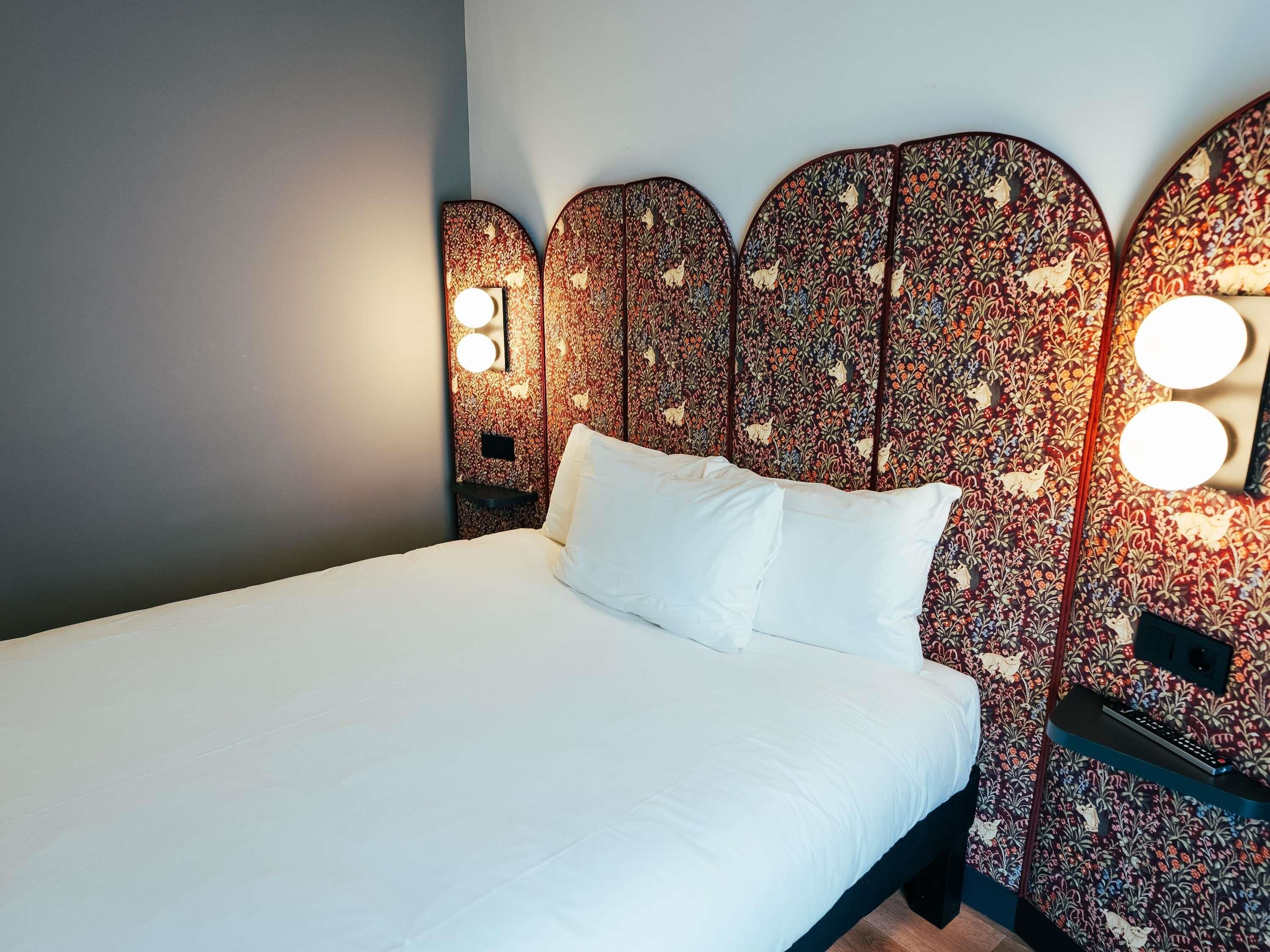 A bed with white linens, a headboard with a floral pattern, and two wall-mounted lights.