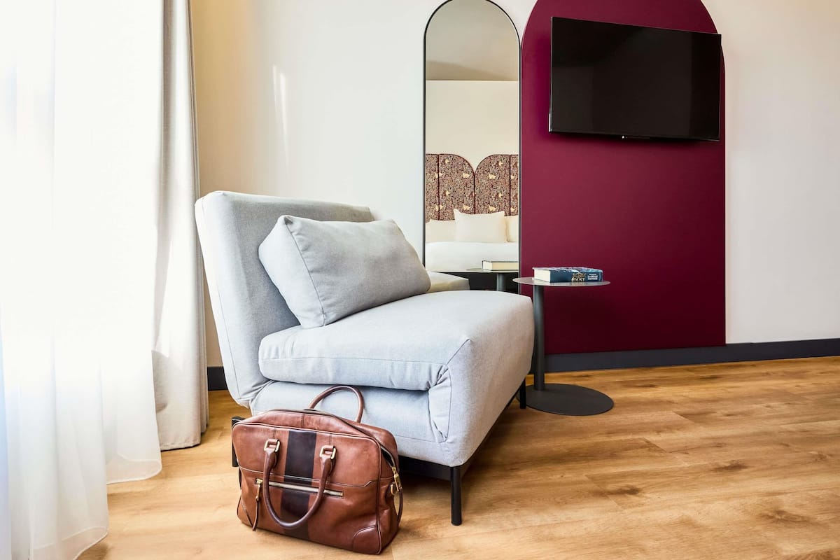 A modern hotel room with a grey sofa, a brown leather bag, a small round table with a book, a mirror, and a TV mounted on a red wall.