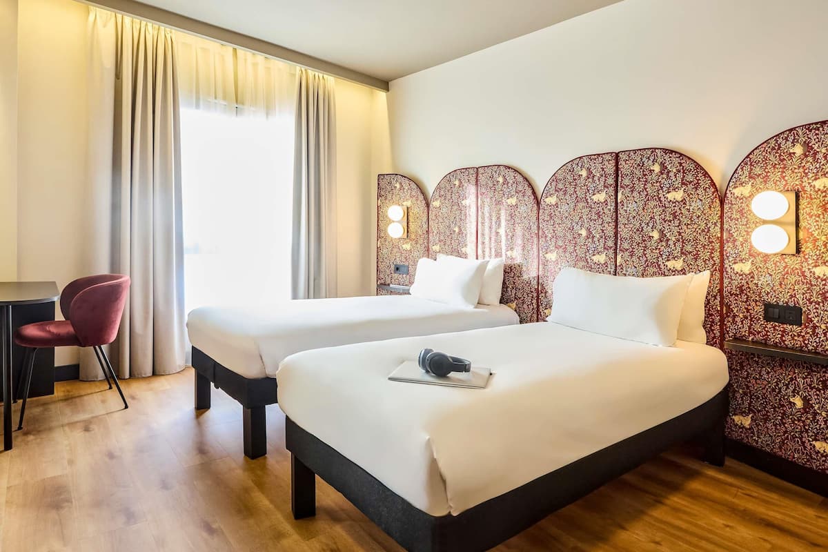 Una habitación de hotel con dos camas, un escritorio, una silla y un ventanal con cortinas.