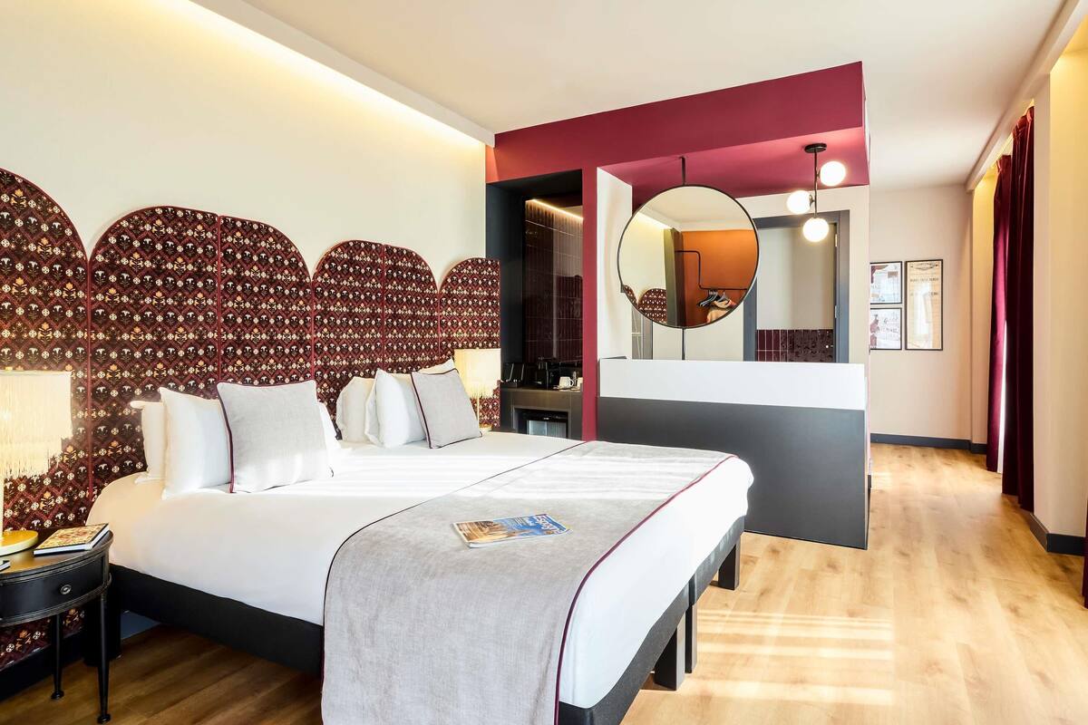 Una habitación de hotel moderna con una cama grande, una mesita de noche, un espejo y vistas al pasillo.