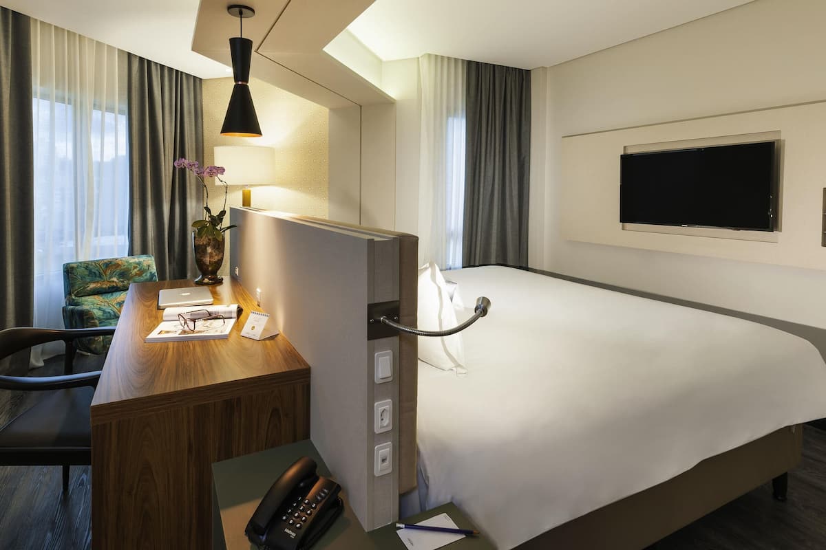 A hotel room with a wooden desk, a telephone, a bed, a television, and a window with curtains.