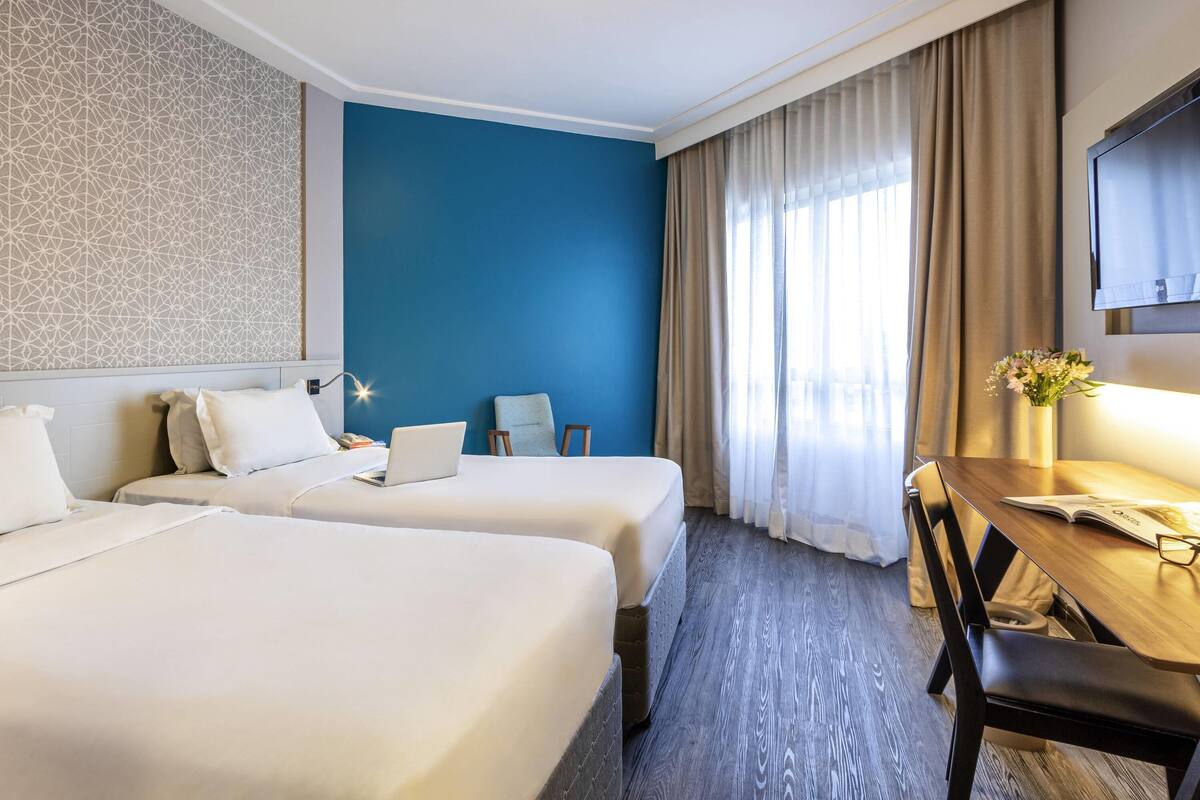 A hotel room with two beds, a desk, a chair, a TV, and a window with curtains.