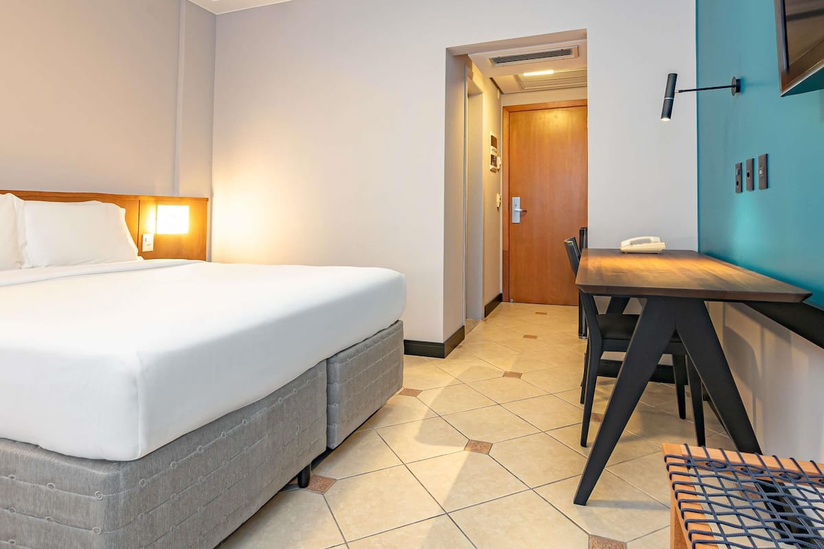 A hotel room with a bed, a desk, a chair, a phone, and a door.