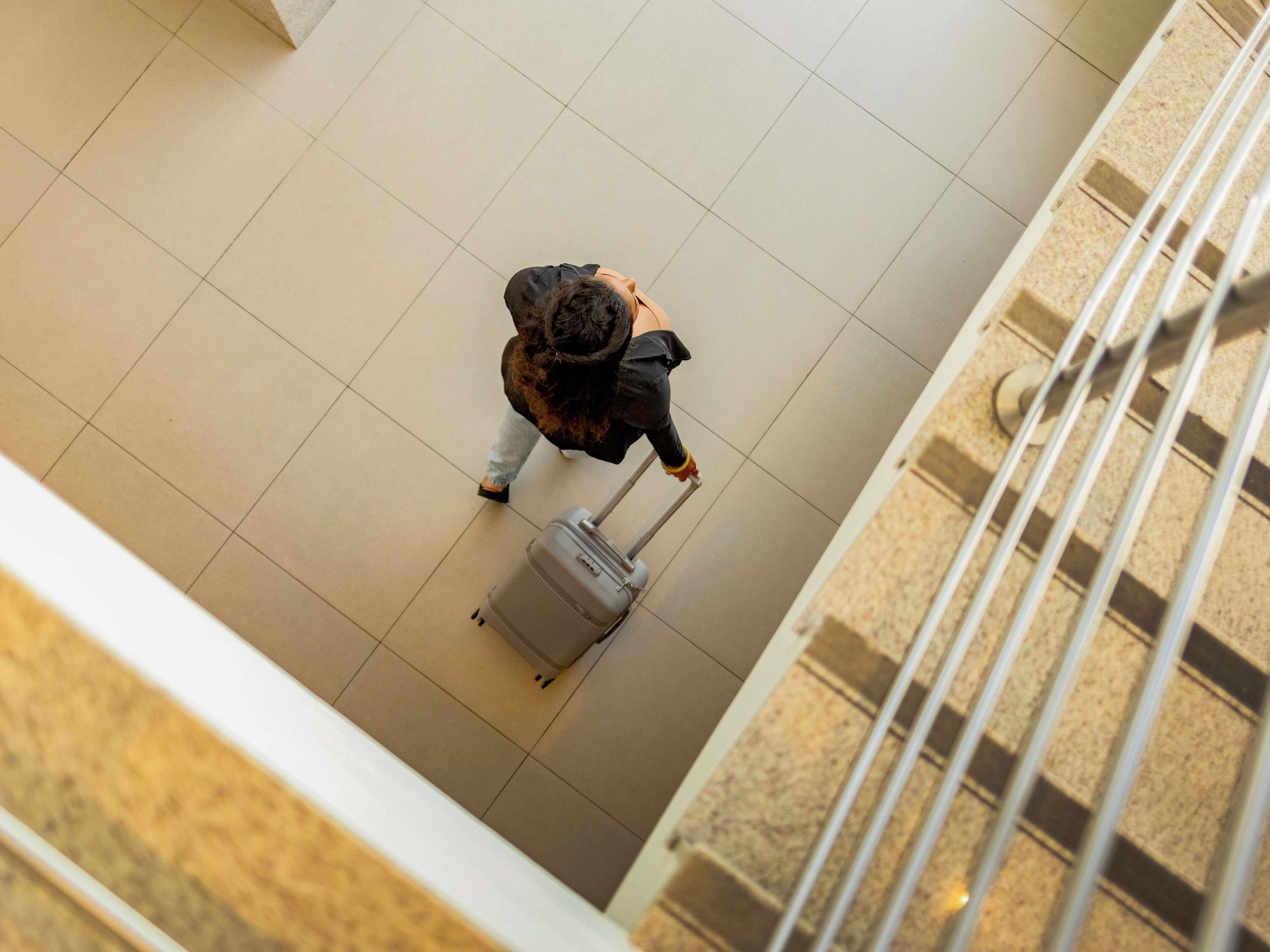 Une personne tenant une valise dans un espace intérieur carrelé, avec une rambarde.