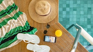 A poolside area with a wooden deck, a striped beach towel, white flip-flops, a straw hat, sunglasses, a yellow cup, and a smartphone on the deck. - Mercure Florianópolis (Florianópolis)