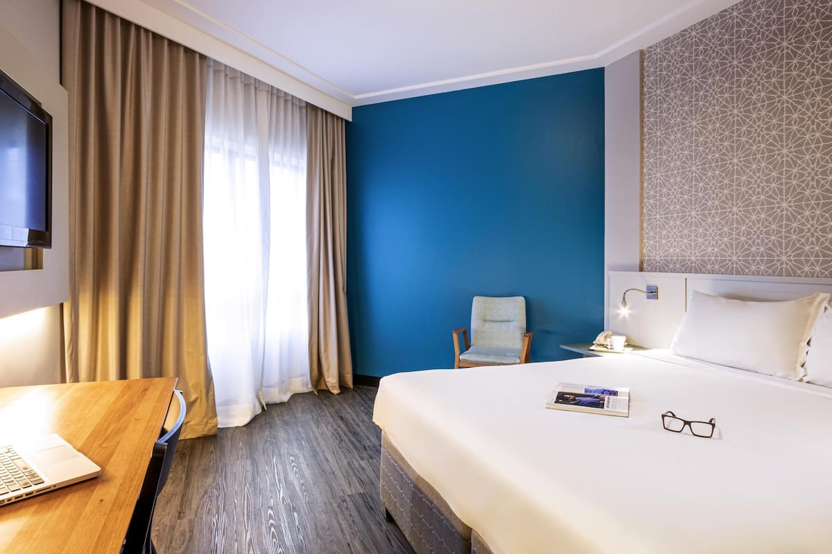 A hotel room with a bed, a desk with a laptop, a chair, and a TV.