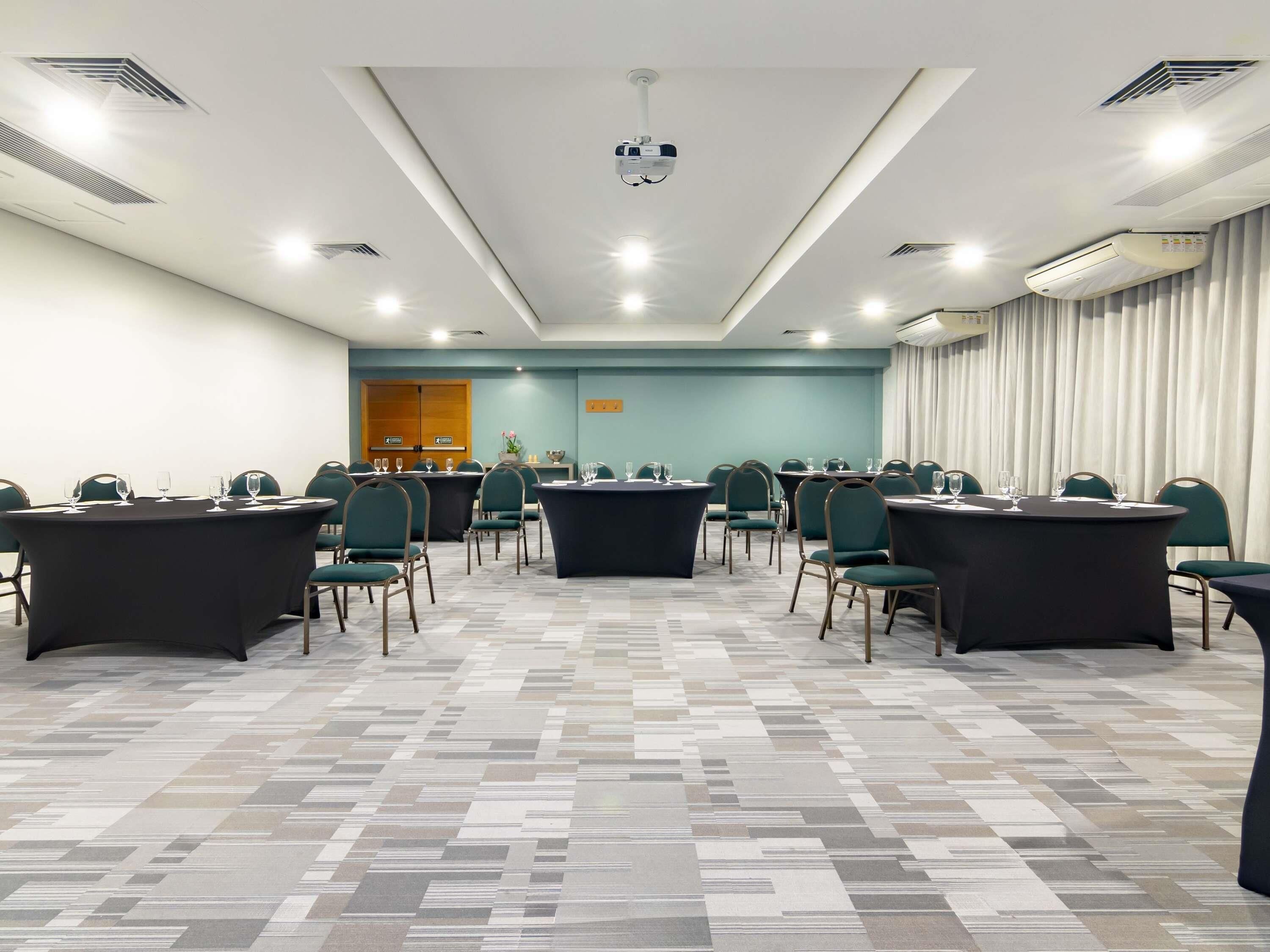 Un salón de conferencias con varias mesas y sillas, un proyector y un ventanal con cortinas.