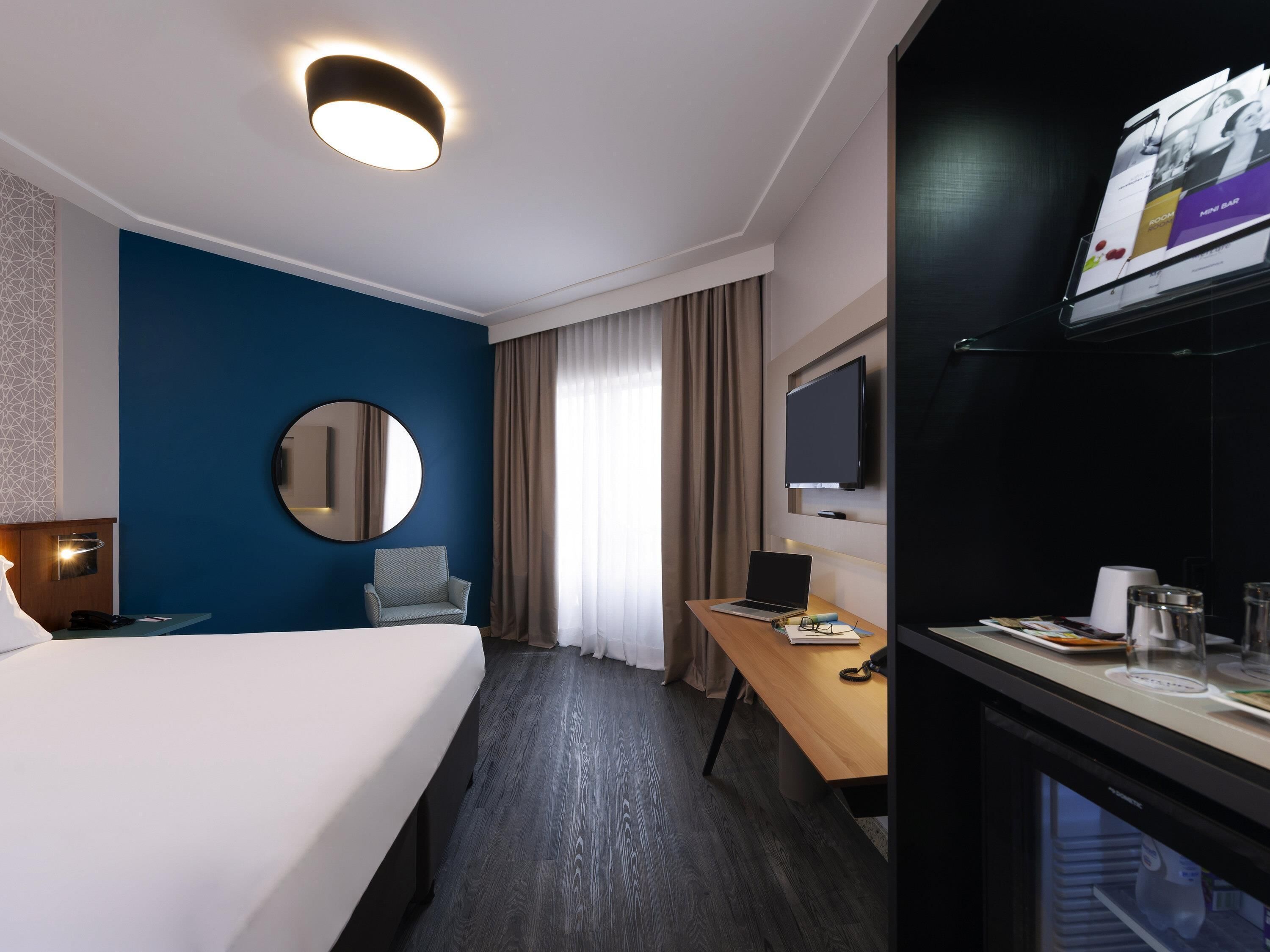 Ett hotellrum med en säng, ett skrivbord med en bärbar dator, en stol, en tv som är monterad på väggen och en minibar.