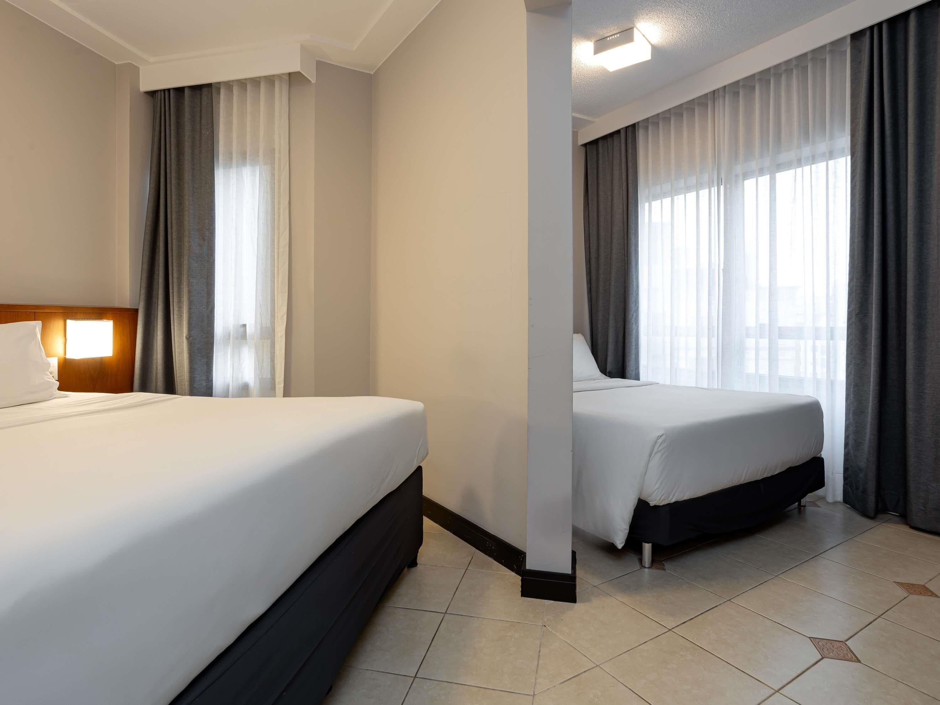 A hotel room with two beds, a nightstand, a lamp, and a window with curtains.