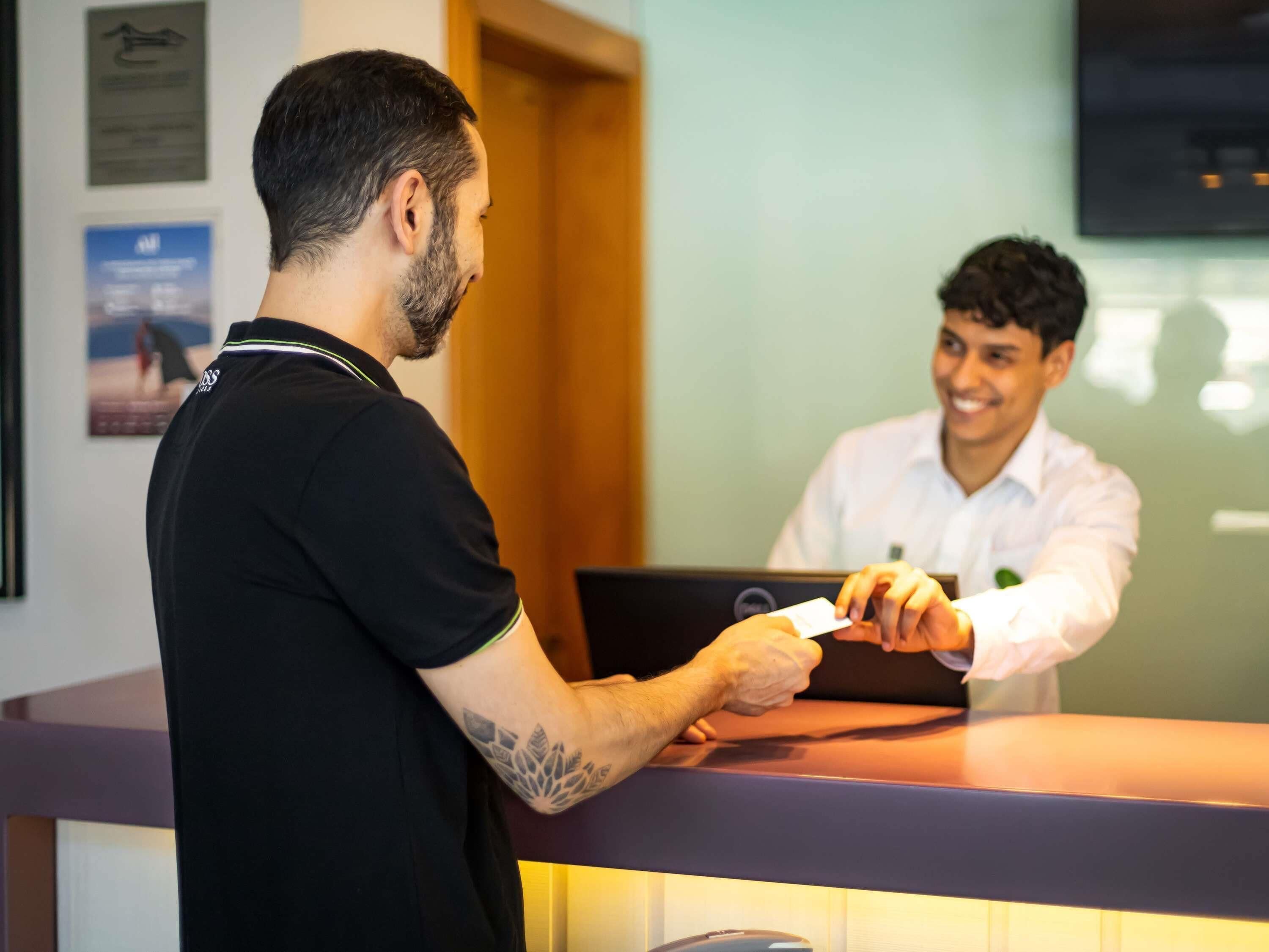 A reception desk with a laptop, a person handing over a card, and a framed certificate on the wall.
