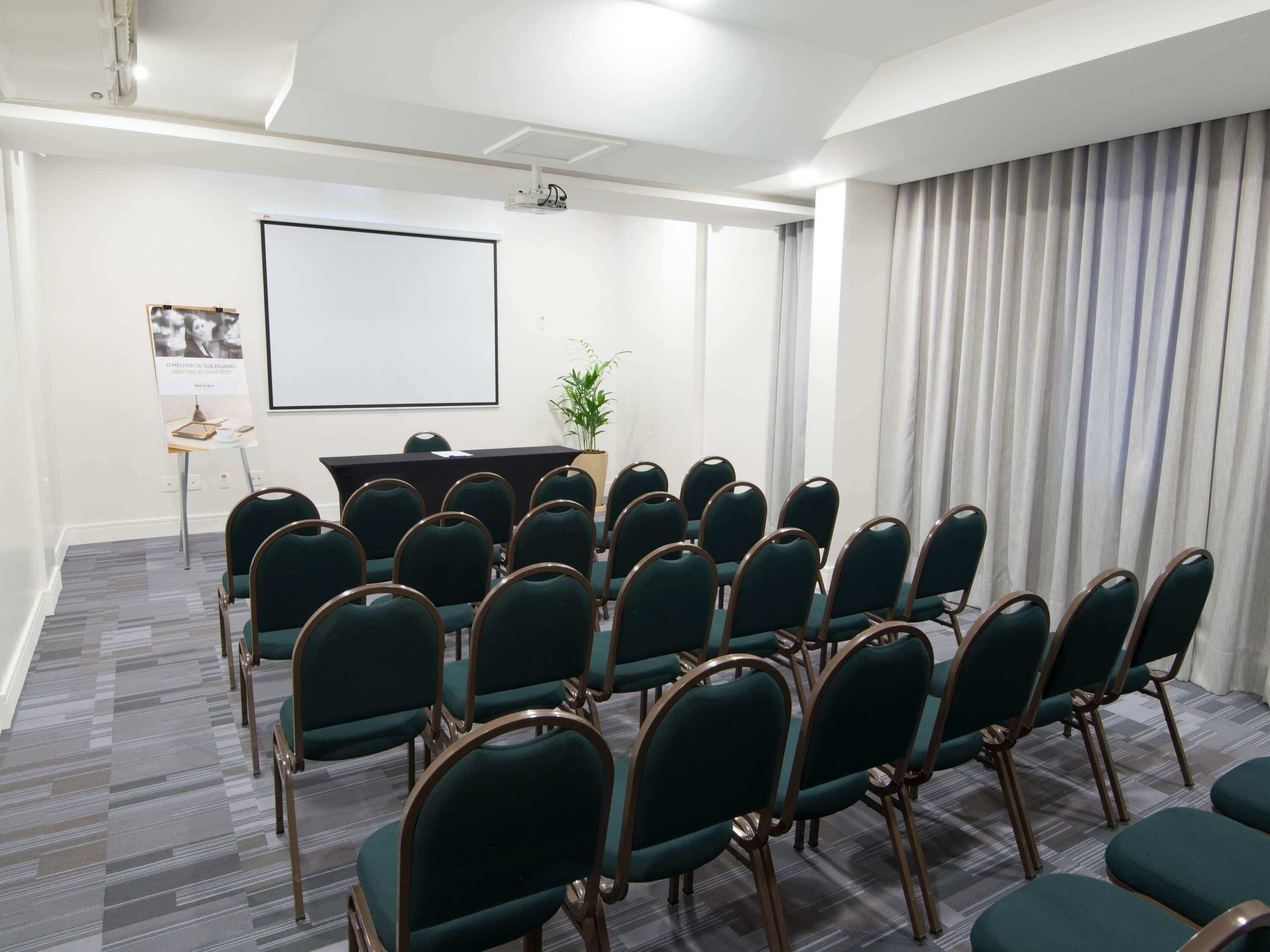 A conference room with rows of green chairs, a projector screen, a whiteboard, and a small table with a plant.