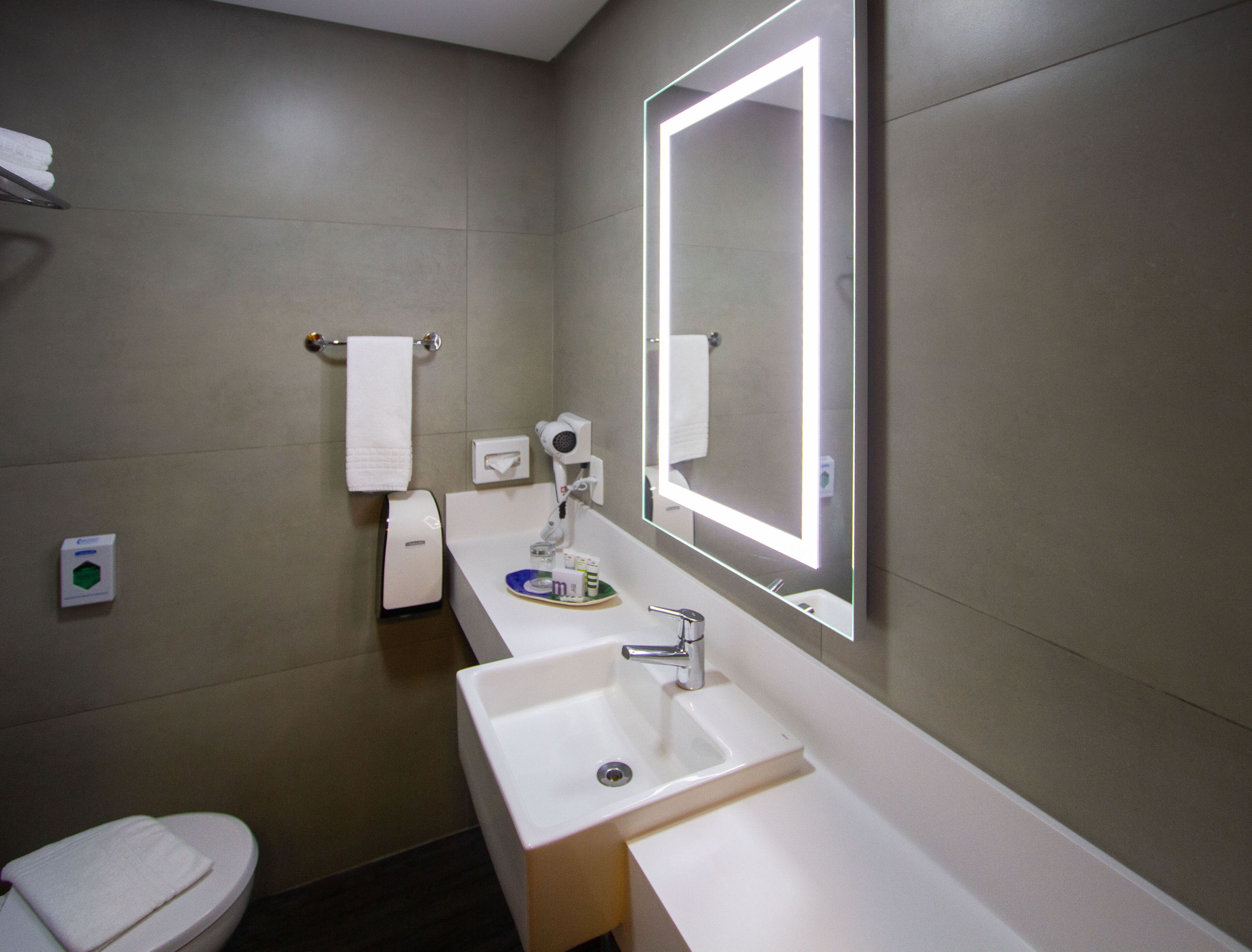A modern bathroom with a toilet, two sinks, a large mirror with backlighting, a towel rack, and a hand dryer.
